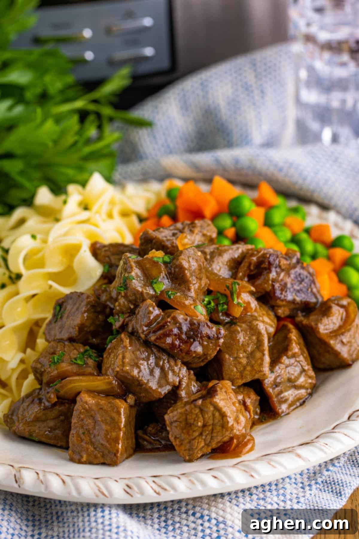 Steak bites, noodles, and vegetables on white plate with glass of water and slow cooker in background.