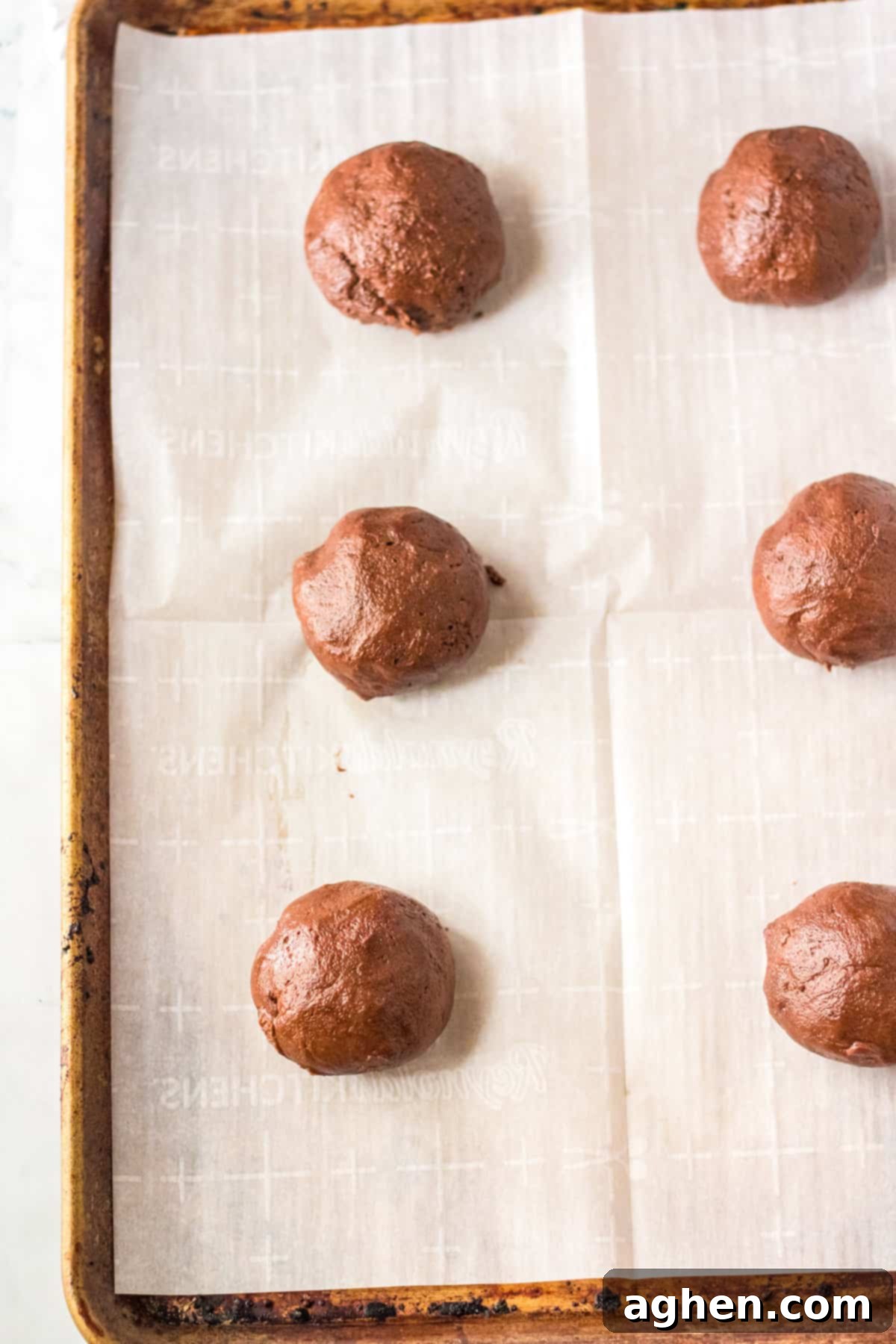 Six large, perfectly rolled chocolate cookie dough balls arranged on a parchment-lined cookie sheet, ready for baking.