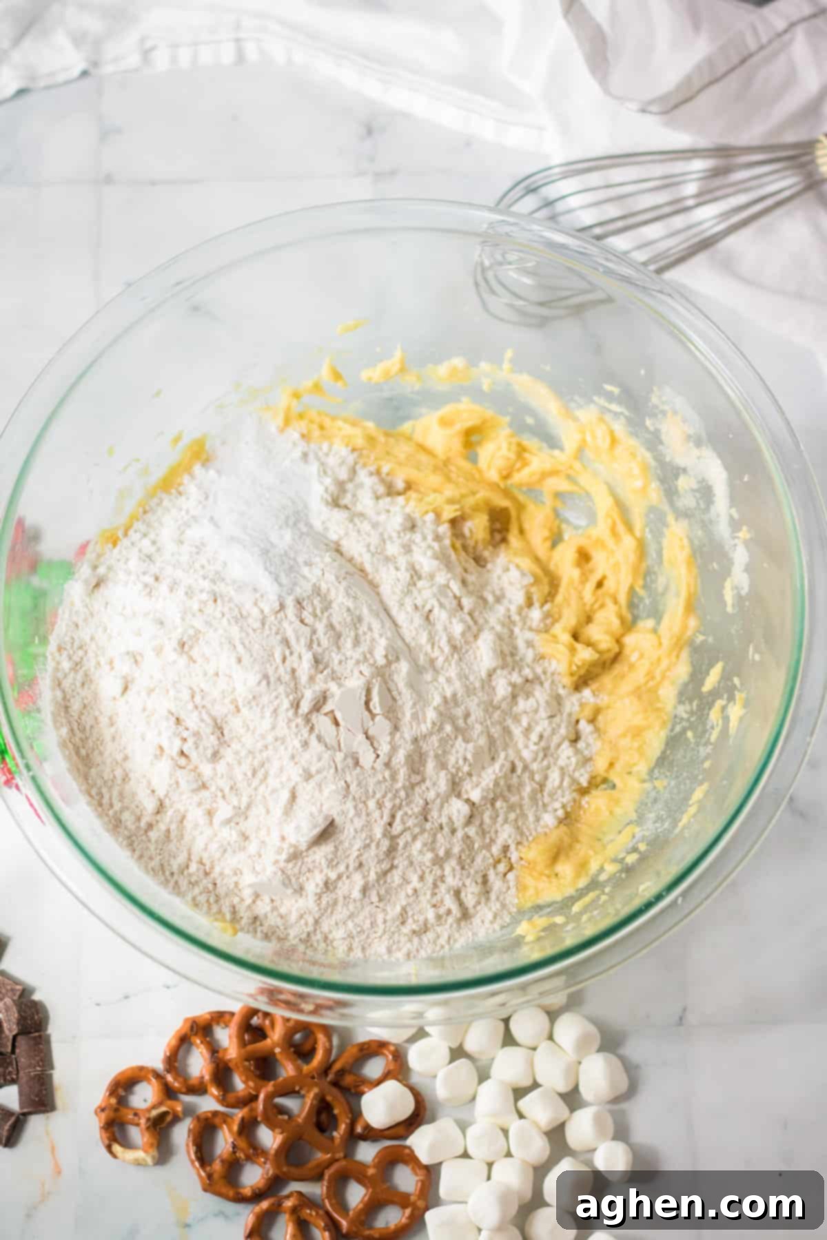 Dry ingredients, including flour, baking soda, and salt, are being carefully added to the wet ingredients in a mixing bowl to form the cookie dough.