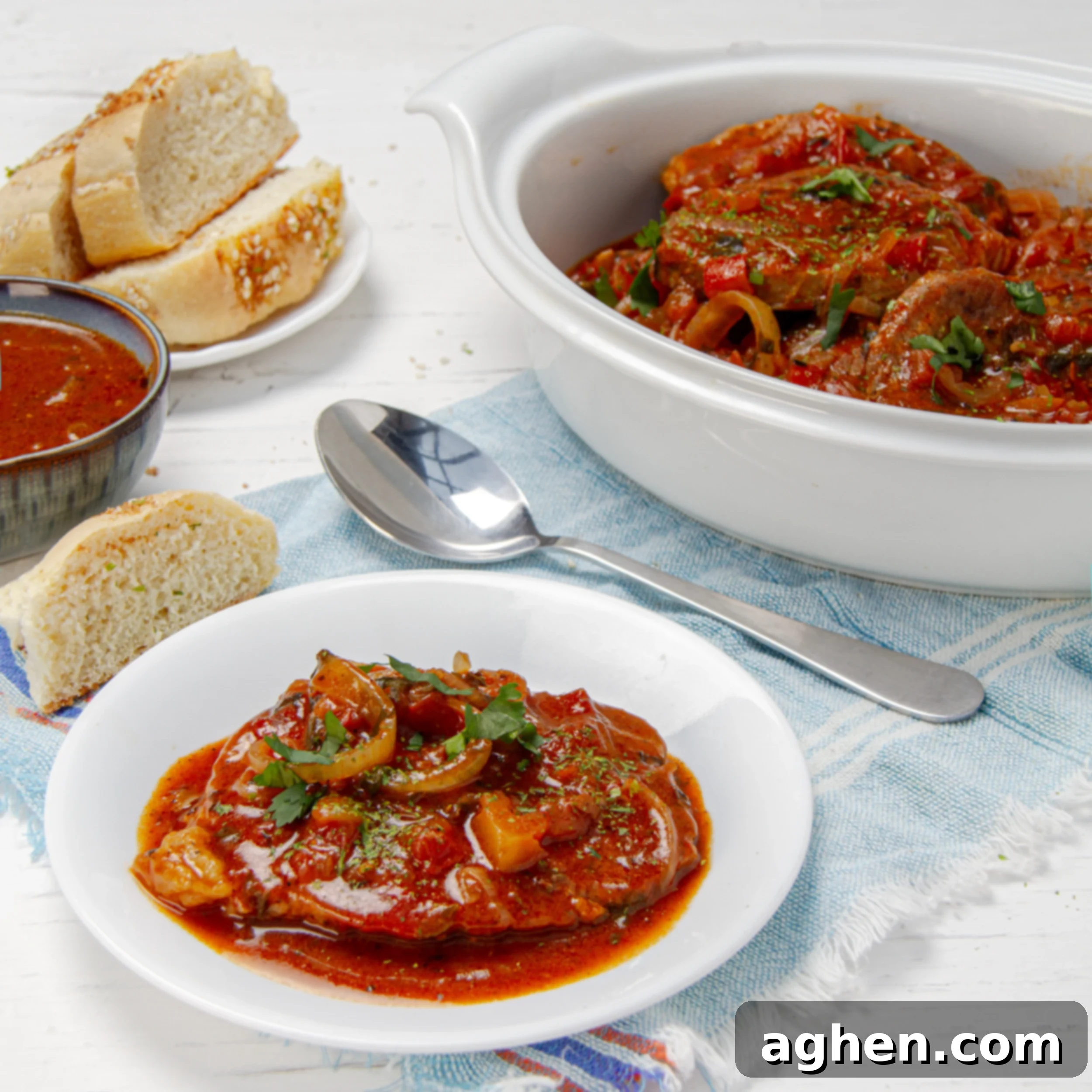 Juicy and tender Swiss Steak in a rich stewed vegetable sauce, presented in an individual serving on a plate alongside the main serving platter with bread and extra sauce.