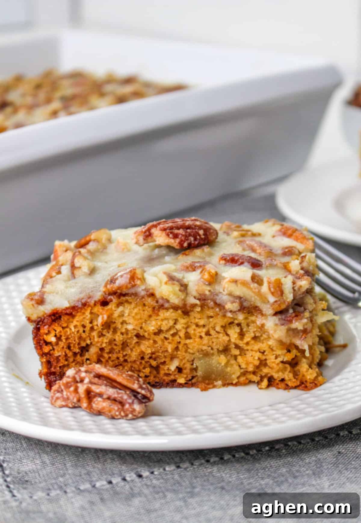 A single slice of fruit cocktail cake on a white plate, elegantly topped with a whole pecan. The texture and rich frosting are clearly visible, promising a delicious bite.
