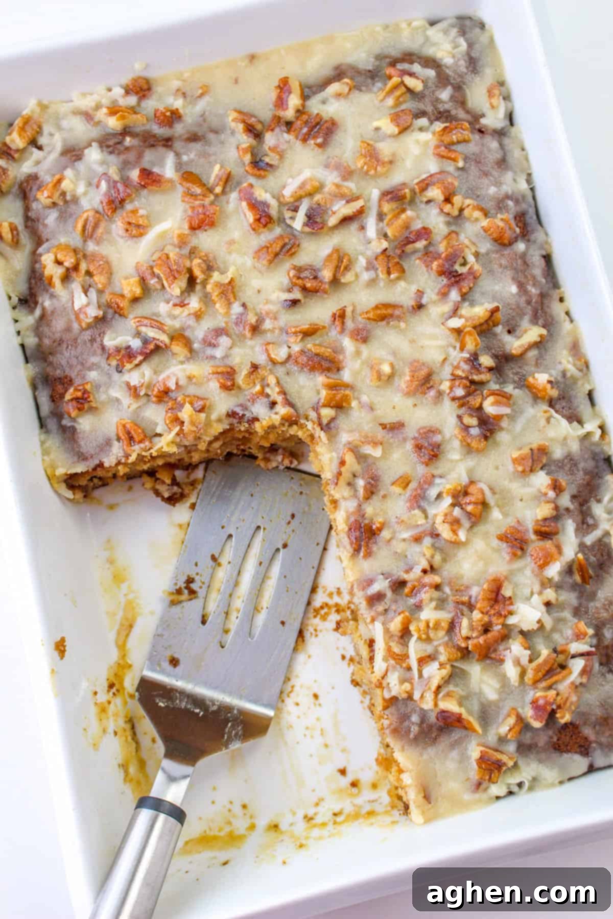A freshly baked fruit cocktail cake, generously covered in a rich coconut pecan frosting, sits in a 9x13 baking dish. A few slices have been removed, with a spatula resting nearby, inviting you to take a piece.