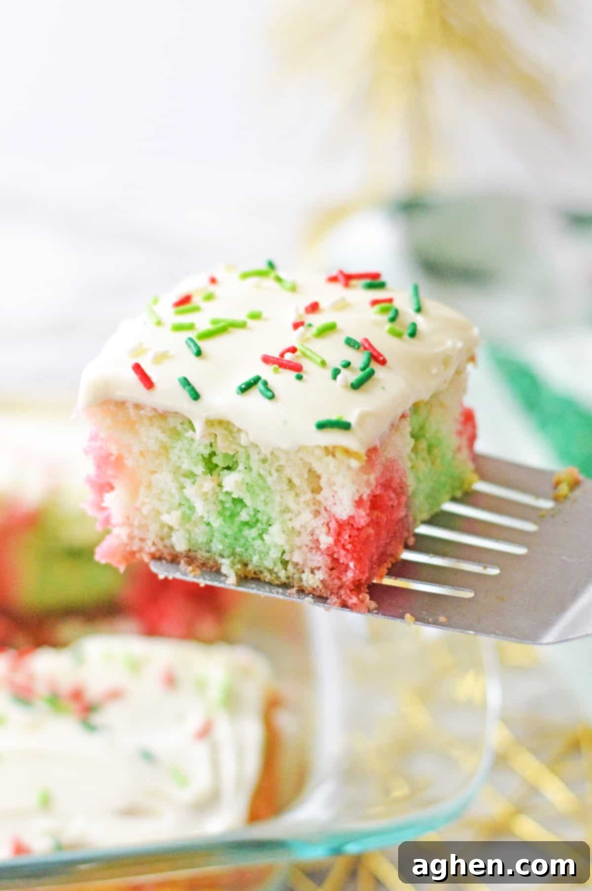 Square slice of red and green jello poke cake being lifted out of baking dish with metal spatula, highlighting the cake's vibrant interior.