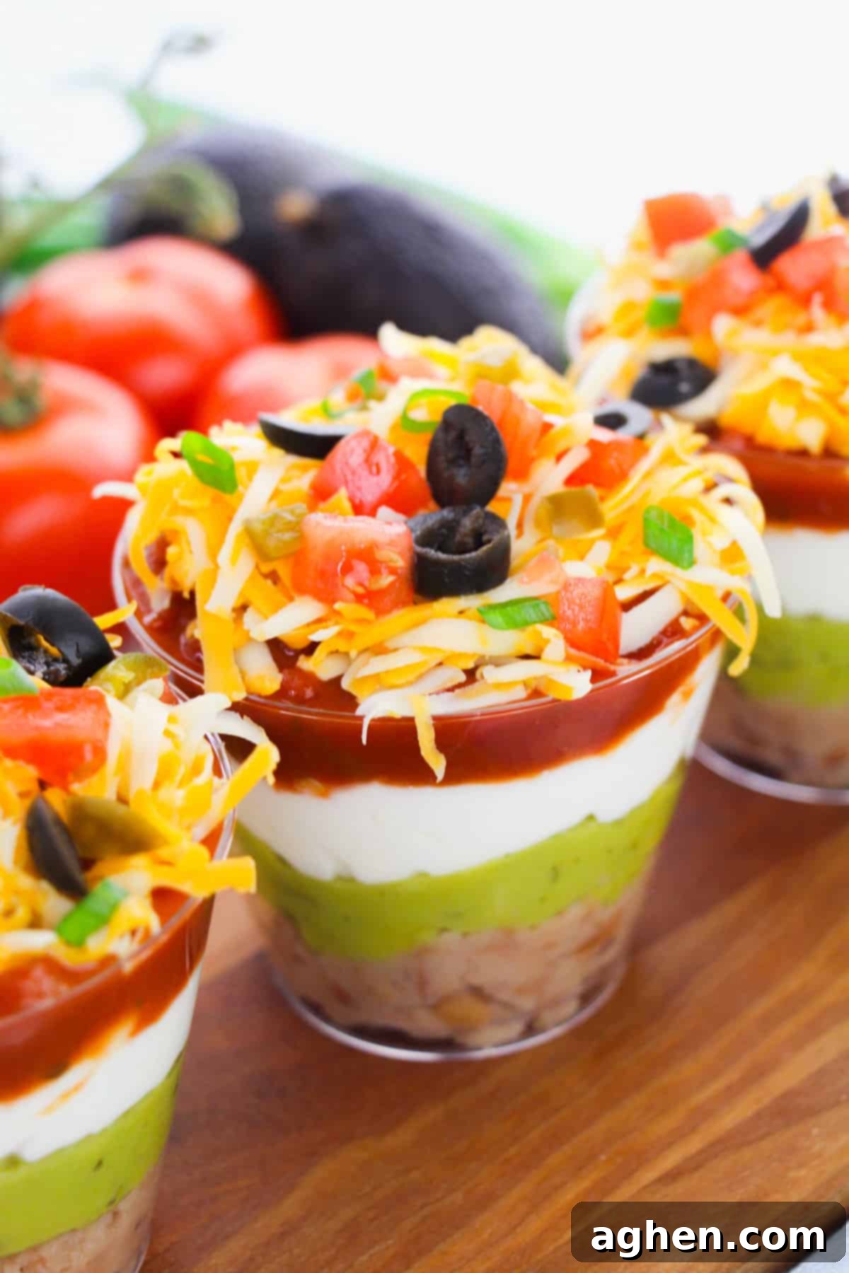 Individual 7 layer dip cups, perfectly portioned for a party. Each clear cup showcases distinct layers of refried beans, guacamole, sour cream, salsa, cheese, and fresh toppings.