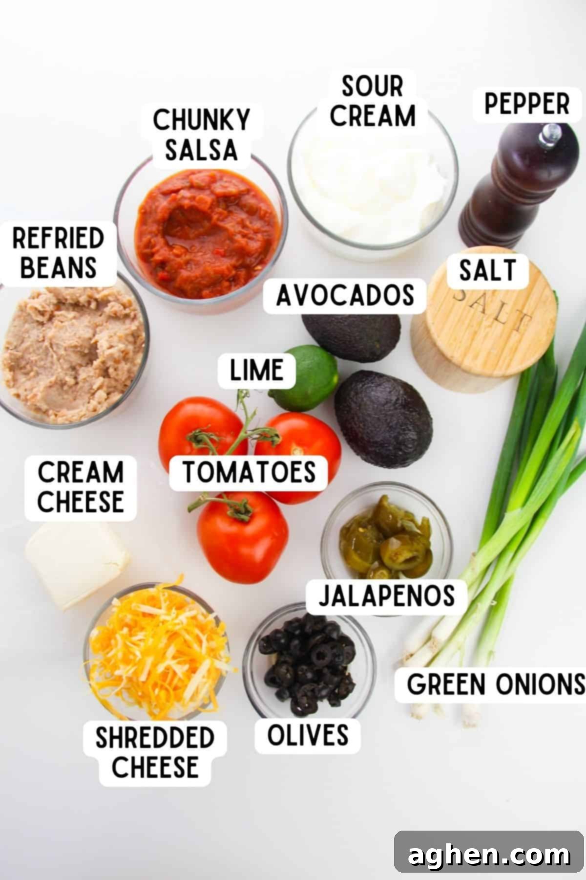 Array of fresh ingredients for 7 layer taco dip, including avocados, refried beans, sour cream, salsa, cheese, and various toppings laid out on a table.