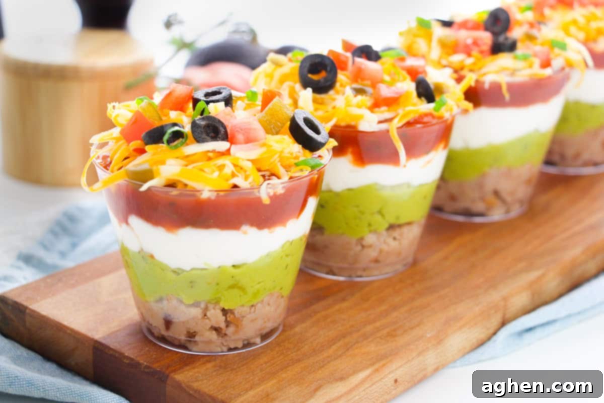 Multiple individual-sized clear cups filled with layered taco dip, showcasing the distinct layers of refried beans, guacamole, sour cream, salsa, and fresh toppings.