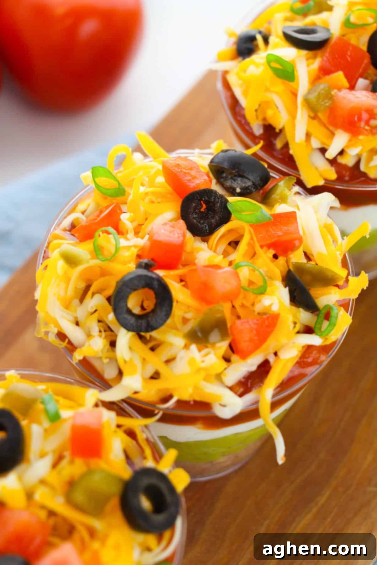 Top-down view of individual 7 layer dip cups, showing vibrant toppings of shredded cheese, diced tomato, sliced black olives, green onions, and diced jalapeños.