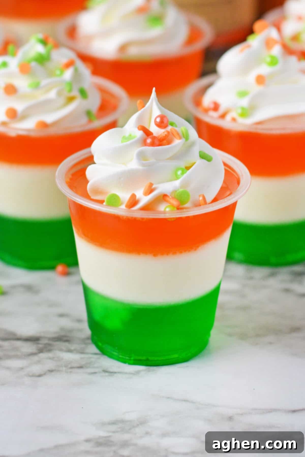 St. Patrick's Day jello shots made with a green bottom layer, white middle layer, and orange top layer, and whipped cream and festive sprinkles, resembling the Irish Flag.