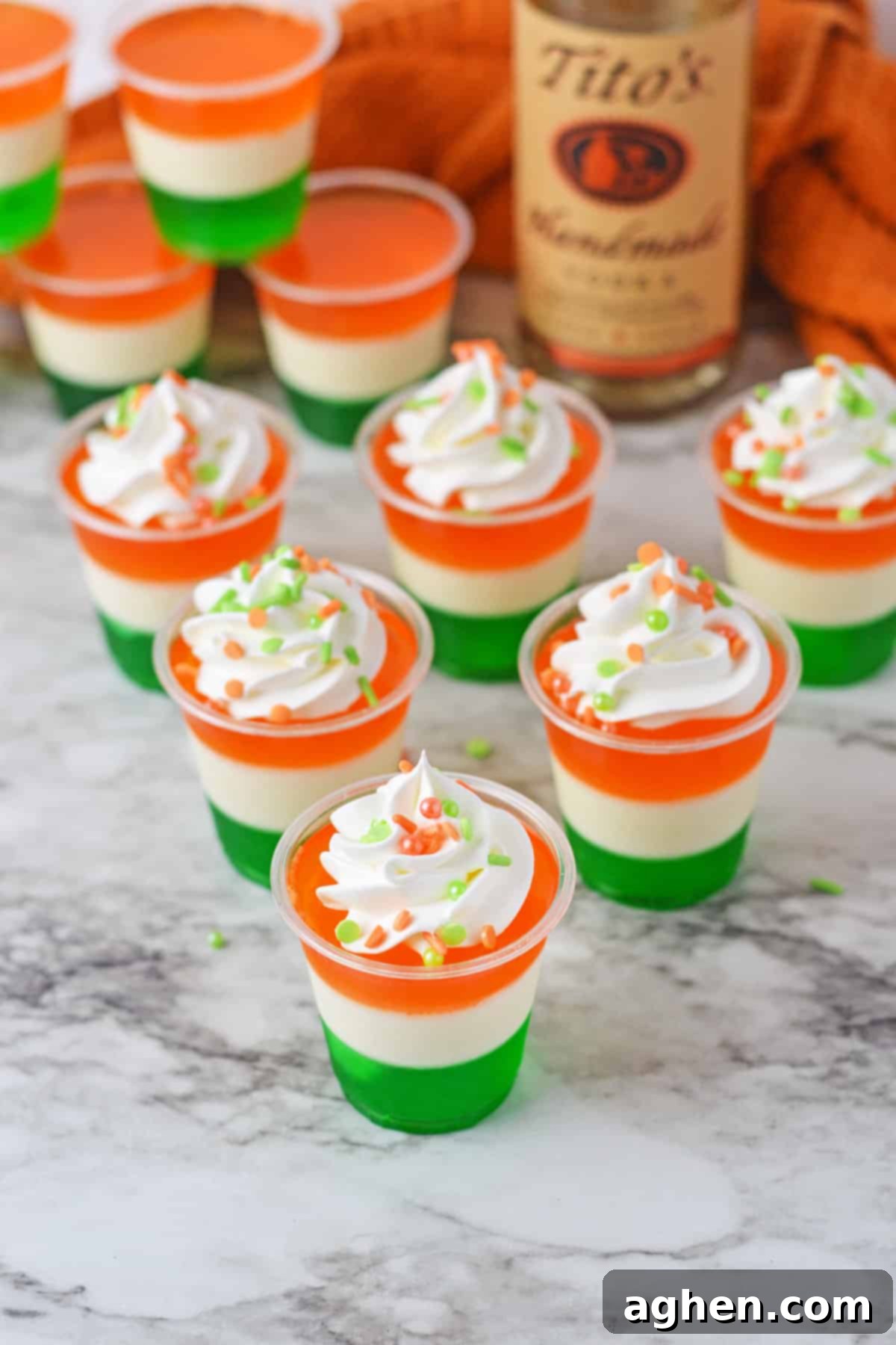 Whipped cream and sprinkle topped Irish flag jello shots with a bottle of Tito's vodka behind them, ready for a St. Patrick's Day party.