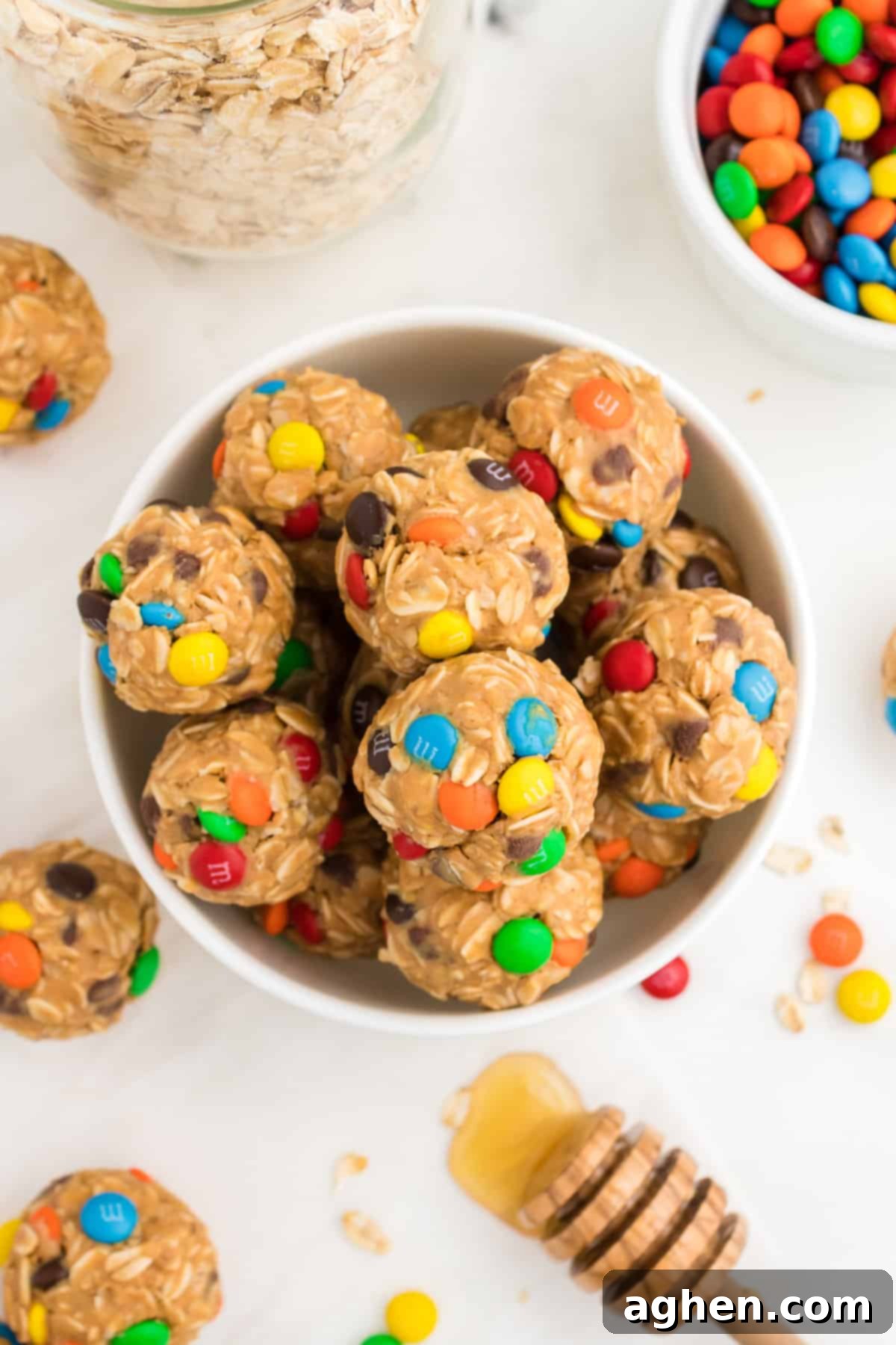 A delightful display featuring a bowl of oatmeal balls with M&Ms, surrounded by a jar of oats, a bottle of honey, a bag of M&M candies, and more finished energy balls.