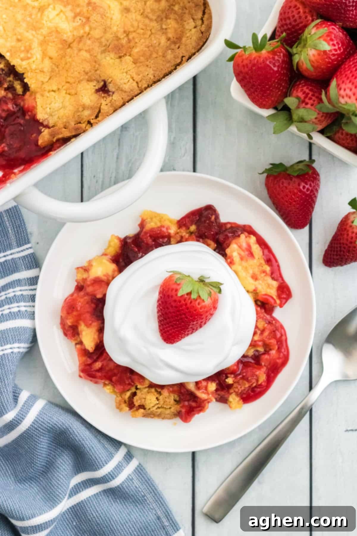A slice of warm Strawberry Dump Cake on a white plate, topped with a swirl of cool whip and a fresh strawberry. In the background, the baking dish holds the remaining cake, a fork, a cloth napkin, and a pint of fresh strawberries, highlighting its deliciousness and ease of serving.
