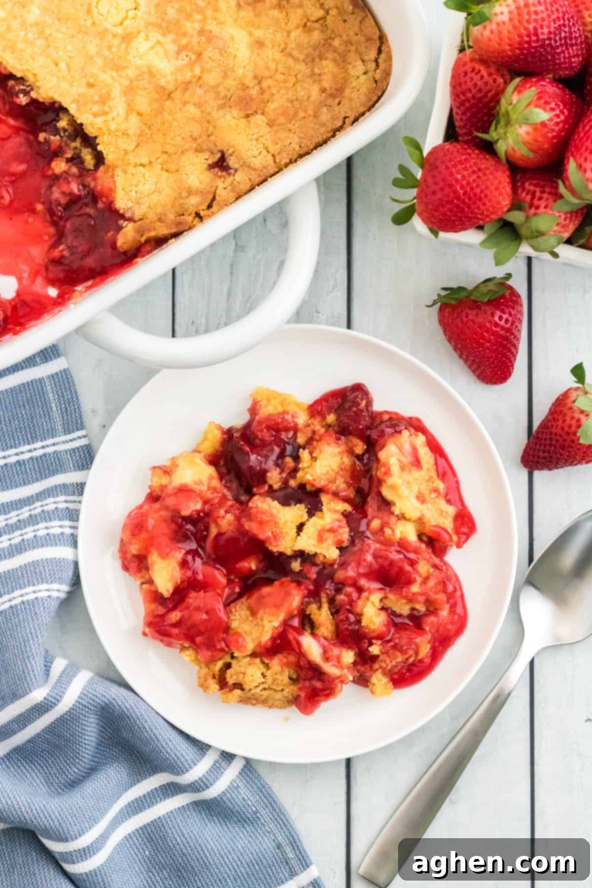 A serving of strawberry pie filling dump cake on a white plate, garnished with fresh strawberries, with the remaining cake in its baking dish visible in the background, inviting diners to enjoy.