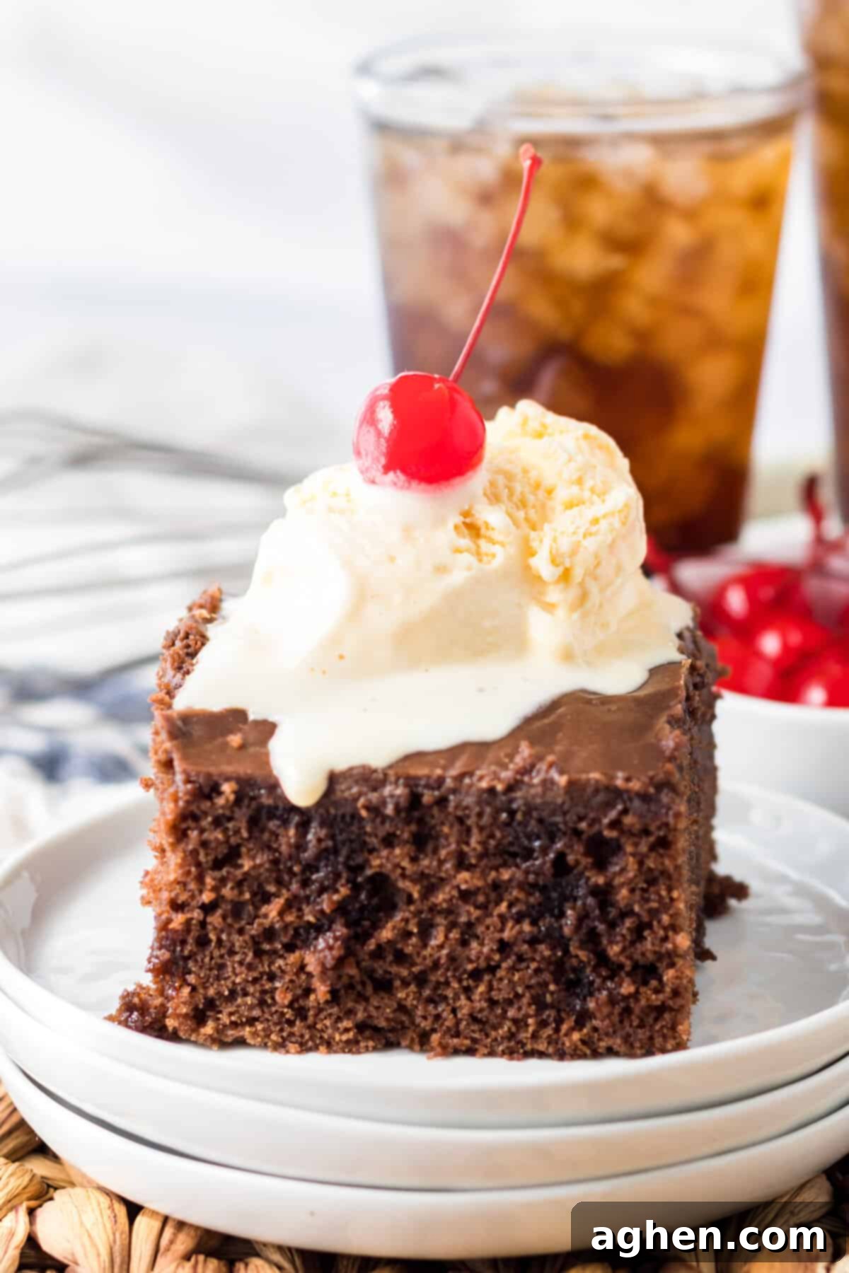 Decadent Double Chocolate Coca Cola Cake 2 A generous slice of double chocolate Coca-Cola cake, beautifully topped with a melting scoop of vanilla ice cream and a vibrant red cherry. A glass of sparkling soda sits elegantly behind the cake, completing the perfect dessert scene.
