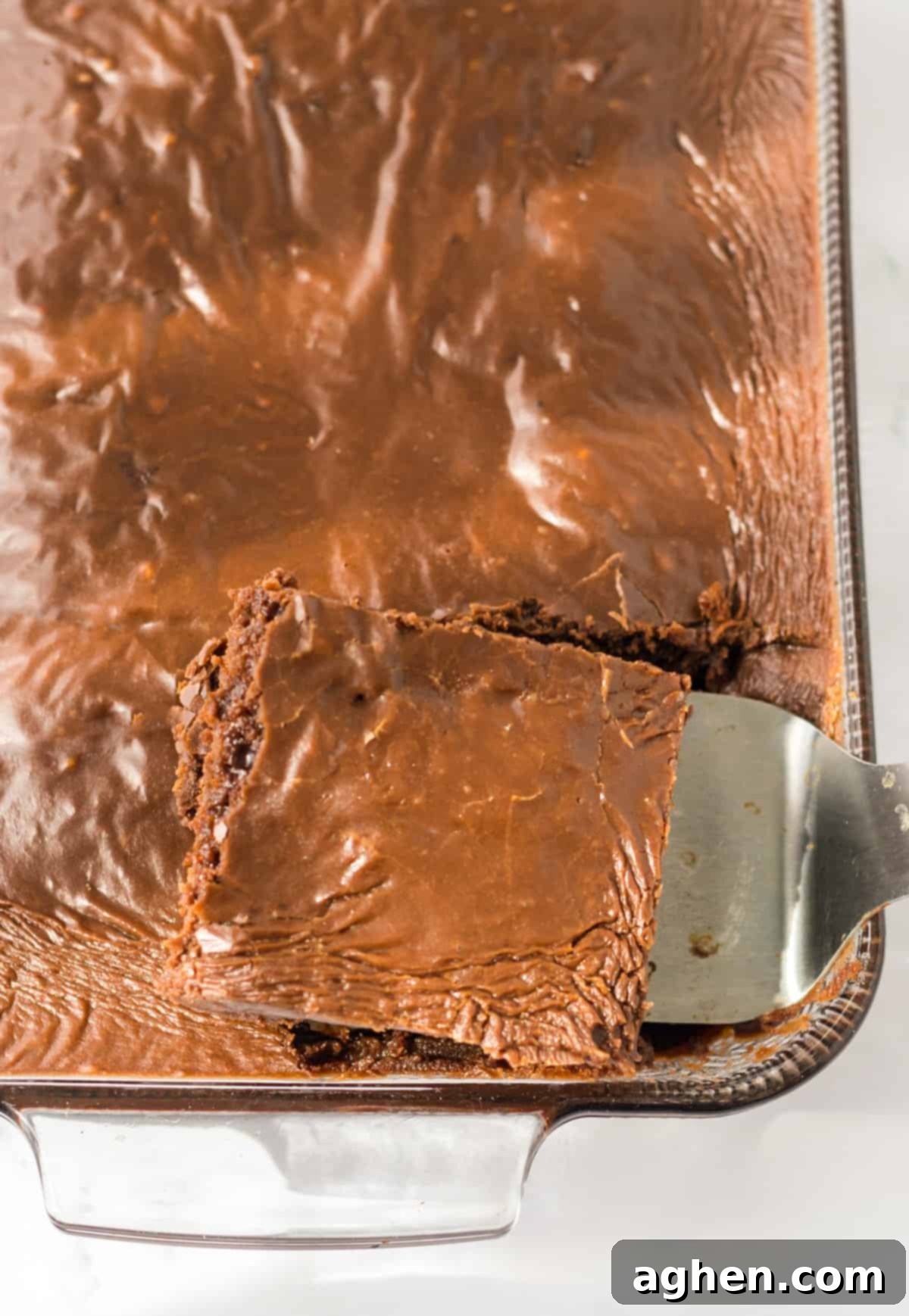 Decadent Double Chocolate Coca Cola Cake 3 A metal spatula gracefully lifting a perfectly frosted piece of moist chocolate Coca-Cola cake from the pan, showcasing its rich, dark brown color and thick, glossy chocolate frosting.