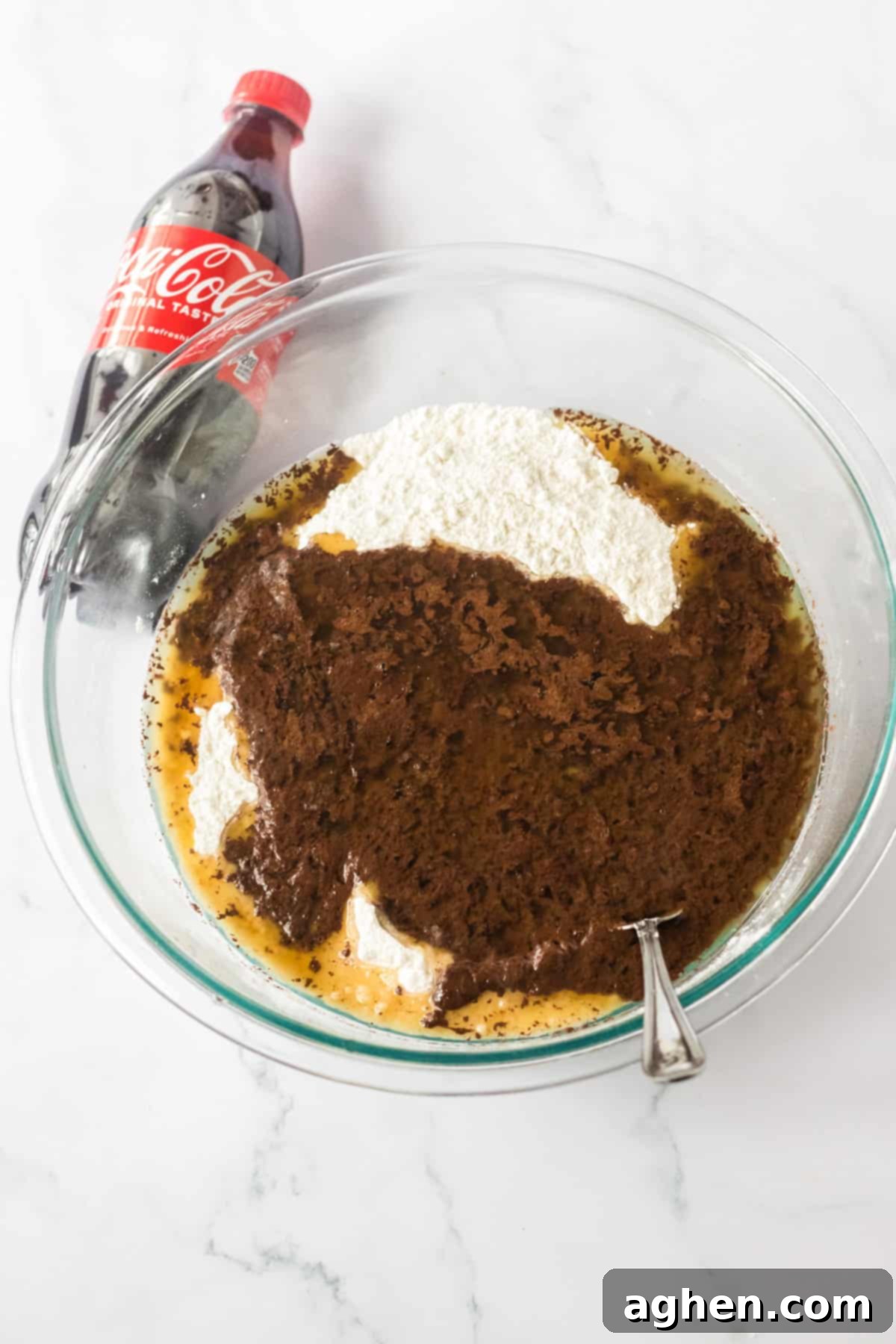 Decadent Double Chocolate Coca Cola Cake 6 A large glass mixing bowl filled with the initial cake batter ingredients, with a spoon leaning on the edge and a bottle of Coca-Cola placed beside the bowl, indicating the next addition.