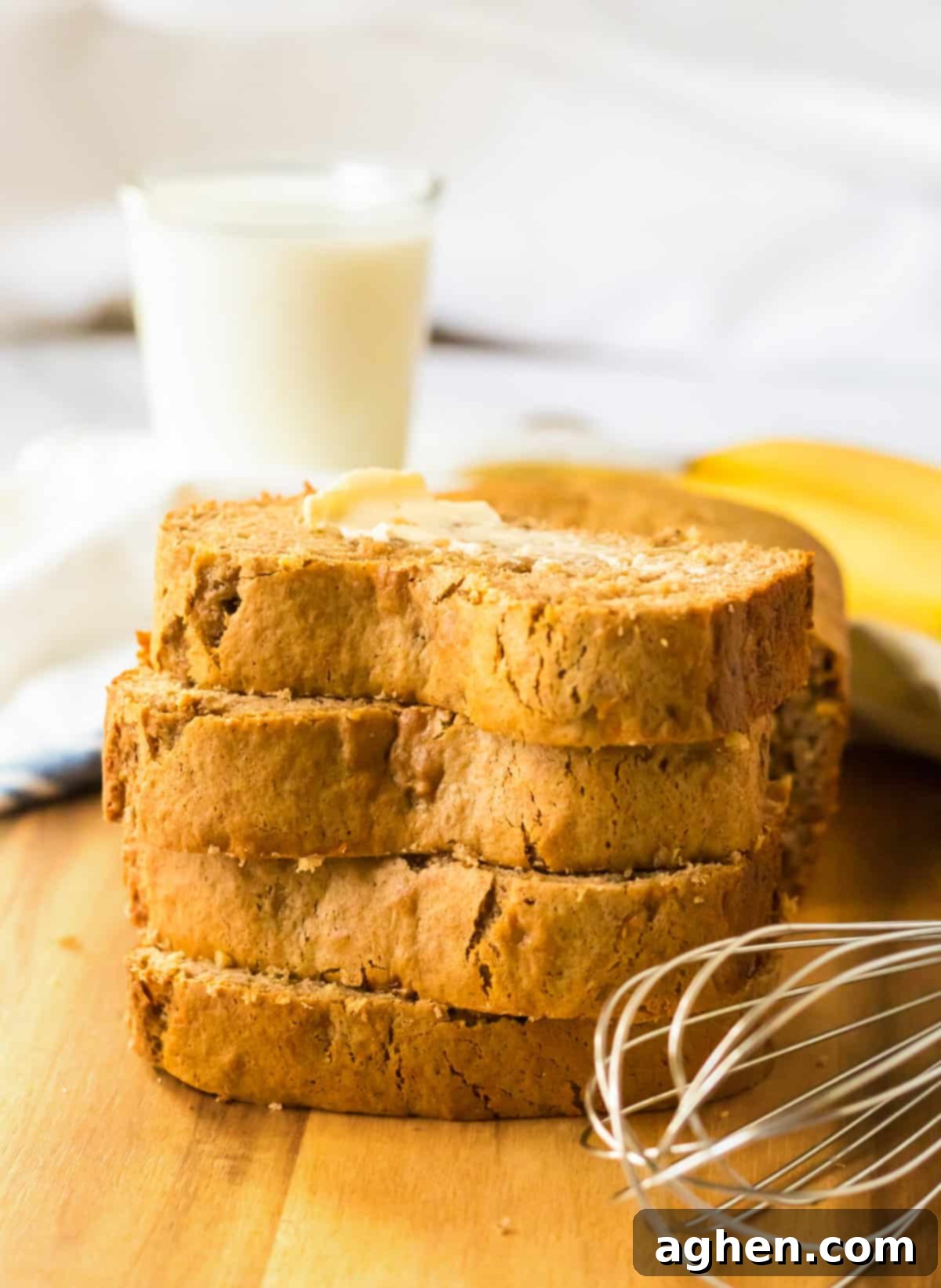 Banana bread slices on a cutting board with a glass of milk, whisk, and bananas.