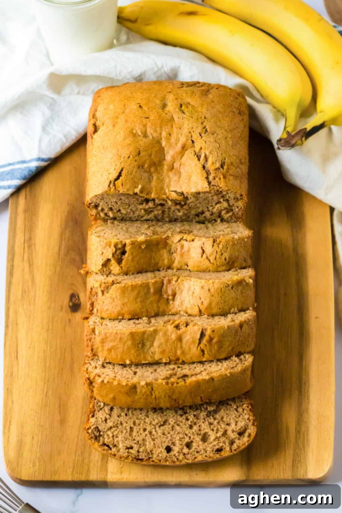 Sliced loaf of banana bread on wood cutting board with a white linen and fresh bananas.