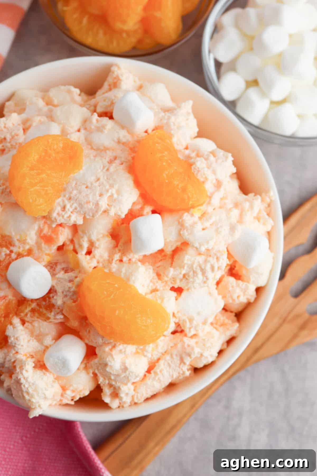 A side view of a serving of fluffy orange jello salad, topped with mandarin oranges and miniature marshmallows, presented beautifully in a clear bowl.