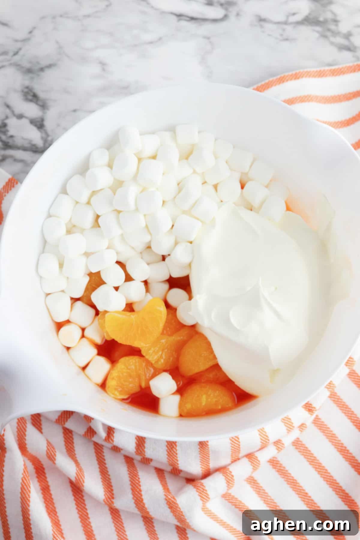 Mandarin oranges, mini marshmallows, and whipped topping are being carefully folded into the chilled orange jello mixture in a large mixing bowl, illustrating the no-cottage-cheese method.