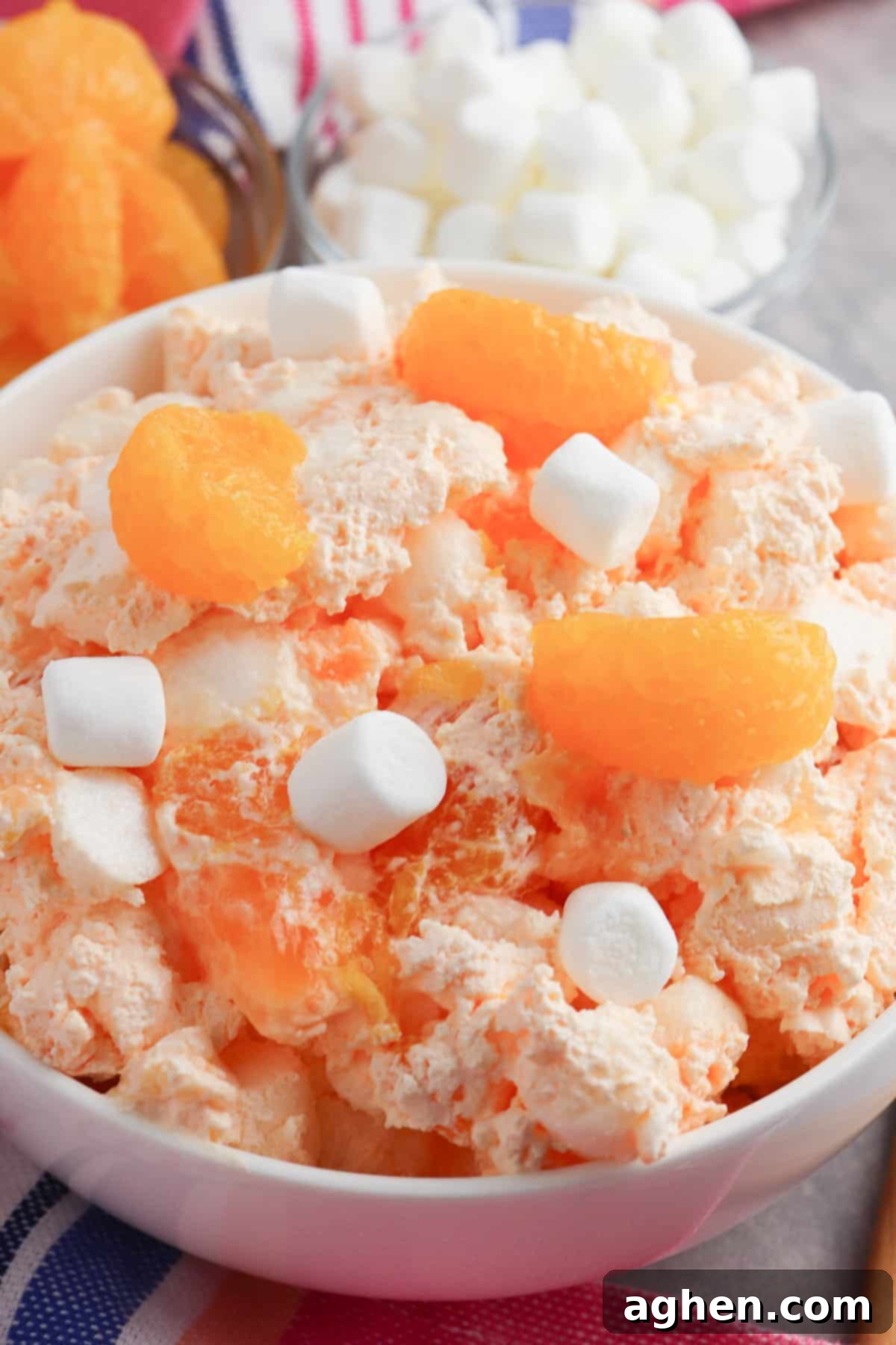 A large glass bowl filled with mandarin orange jello salad, generously mixed with marshmallows, sitting on a rustic wooden surface.