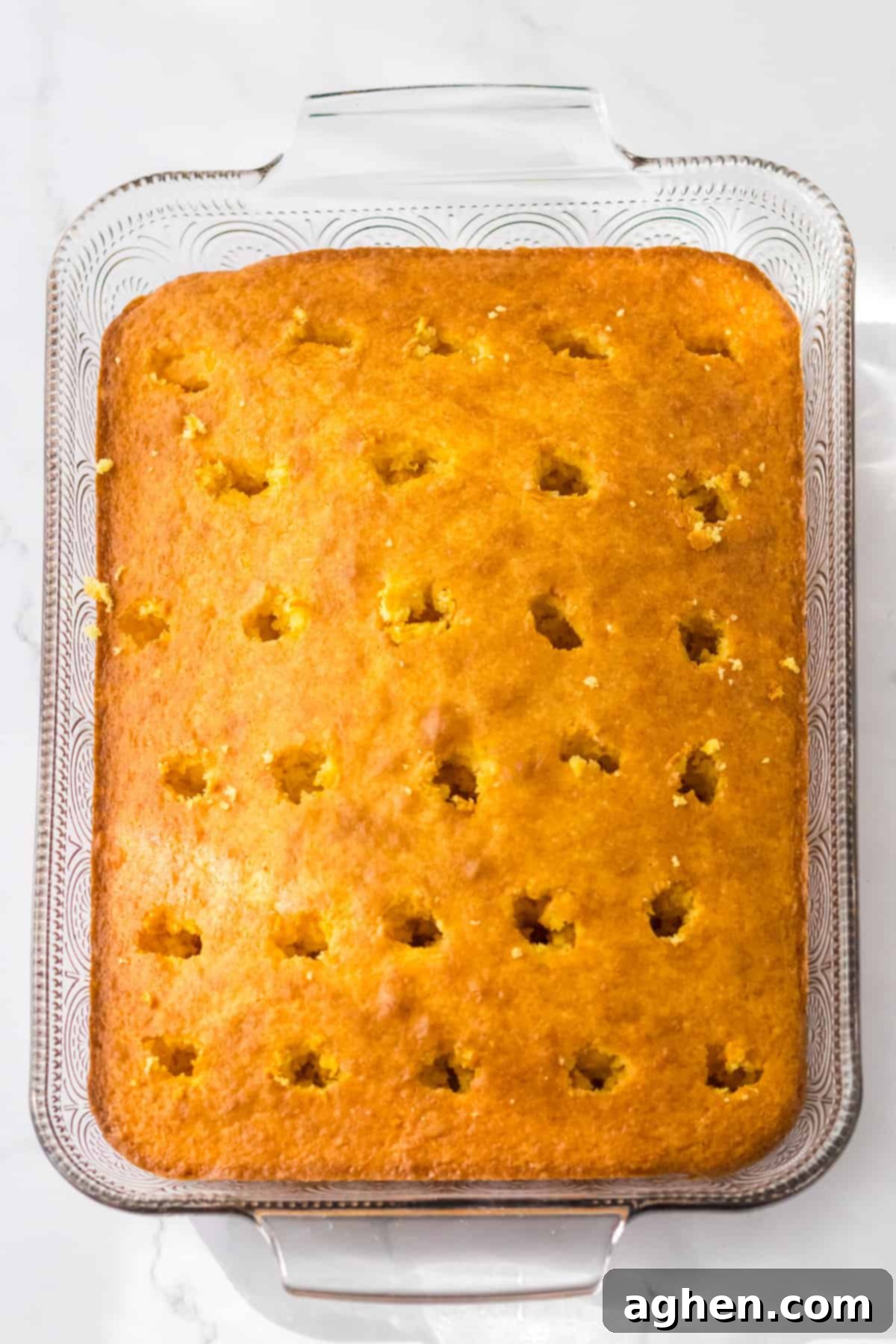 Banana Cream Dream Poke Cake 5 A rectangular yellow cake, freshly baked with a golden-brown top, now features numerous deep, evenly spaced holes created by a wooden spoon, ready to absorb the creamy banana pudding filling.