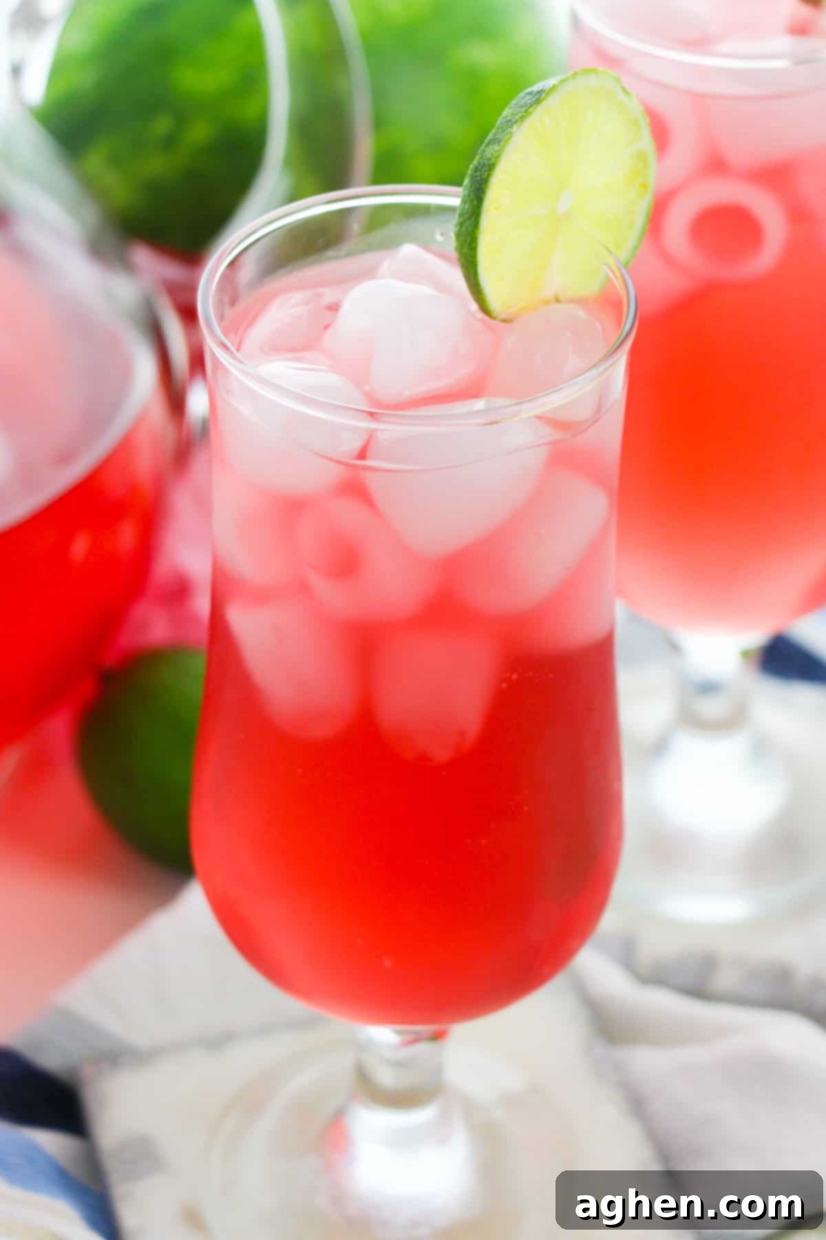 Watermelon refresher drink in glass with ice and lime wheel garnish. A pitcher and another glass of the drink are in background,