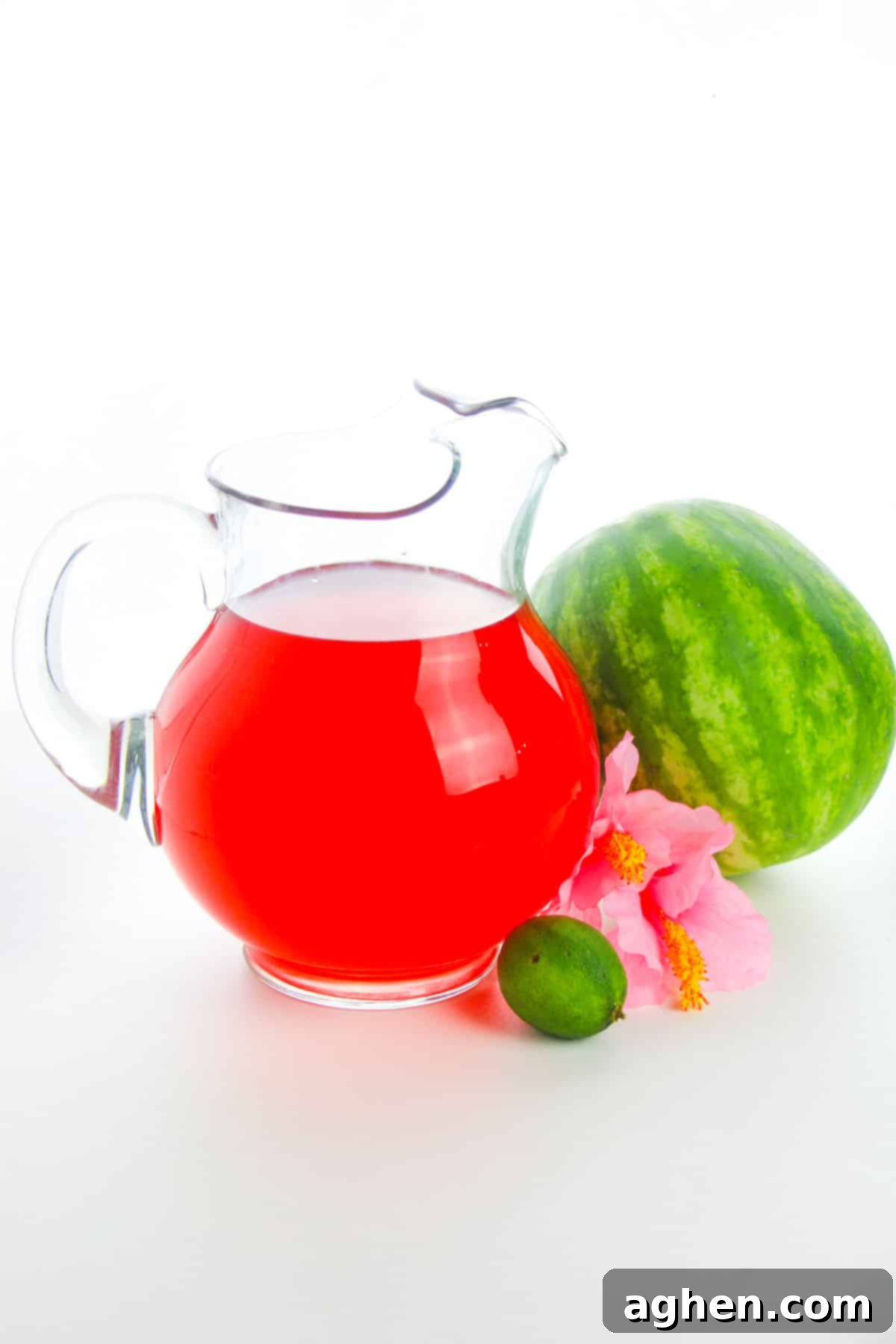 Watermelon drink in large glass pitcher with a small watermelon, a lime, and a pink flower next to it.