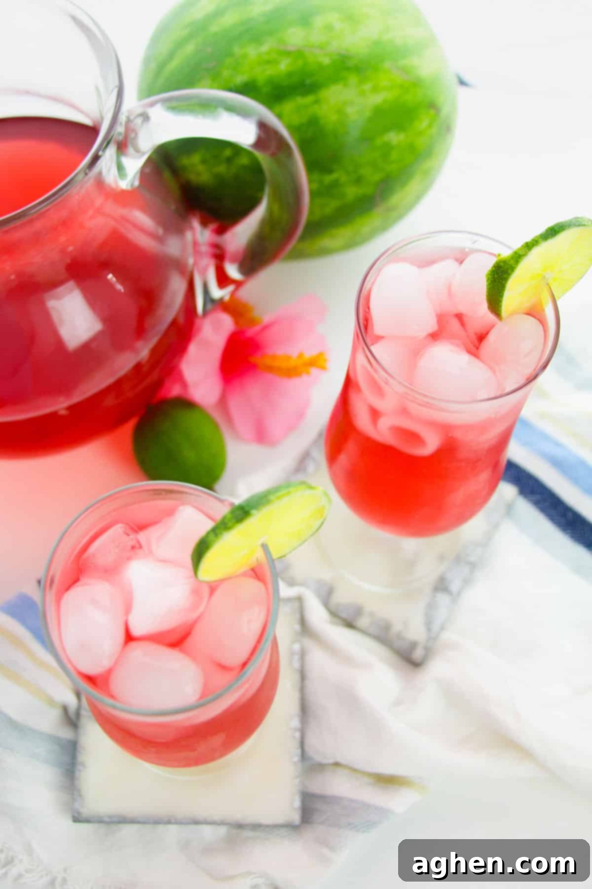 Two watermelon drinks garnished with lime next to watermelon and pitcher.