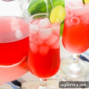 Watermelon refresher drink in glass with ice and lime wheel garnish. A pitcher and another glass of the drink are in background,