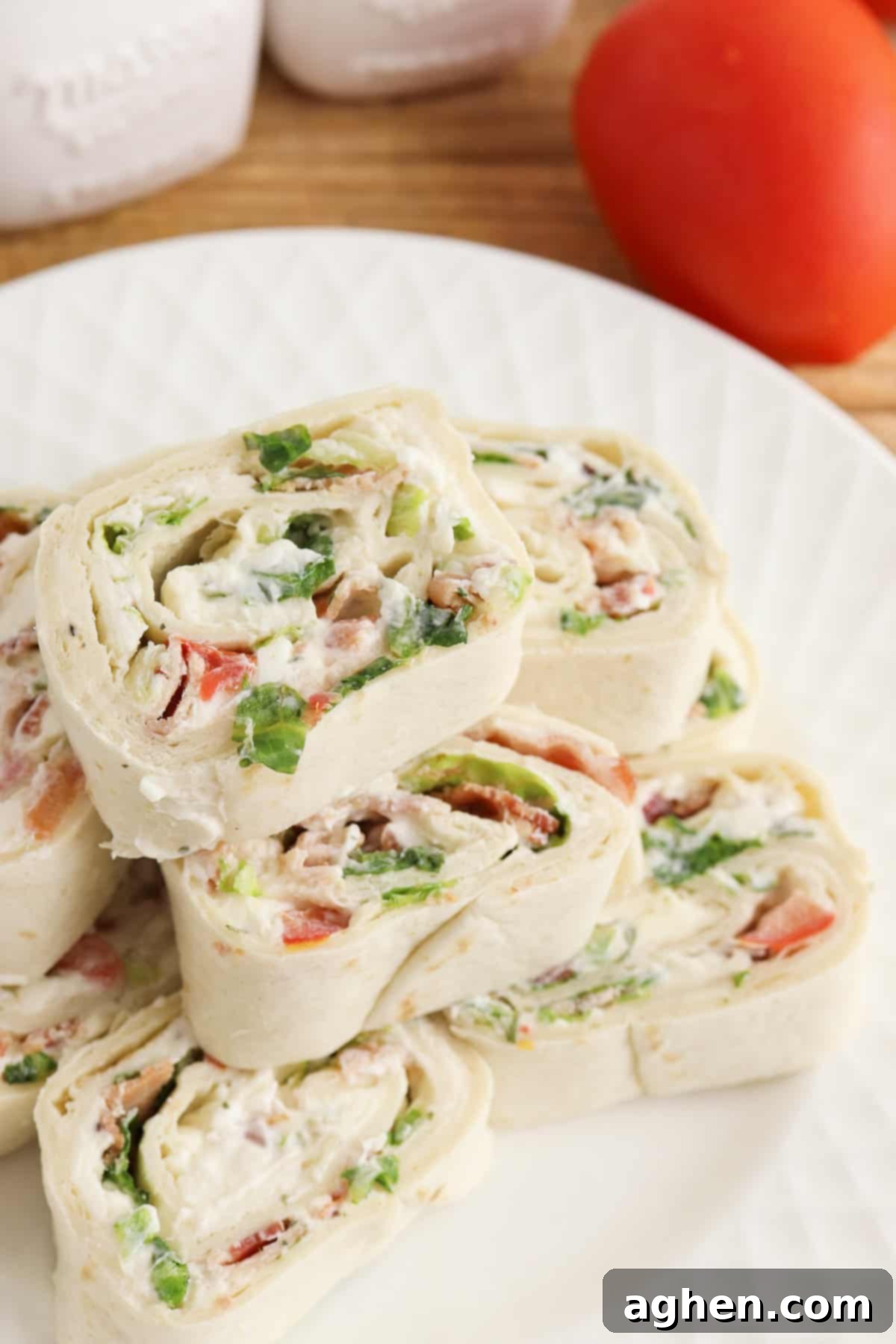 Creamy Ranch BLT Swirls 7 Ranch BLT Pinwheels made with flour tortillas, bacon, lettuce, tomatoes, and ranch dressing stacked on a white plate with a plum tomato next to it.