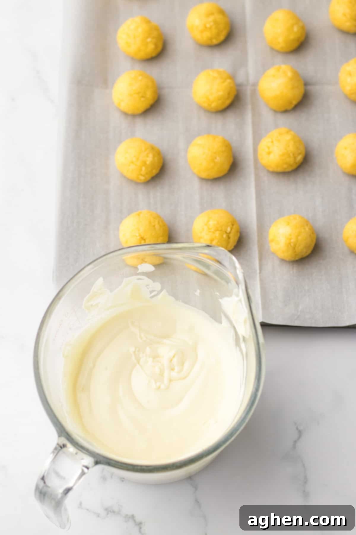 A glass measuring cup filled with perfectly melted white chocolate, with a tray of dipped Oreo truffles visible in the background, set on lined parchment paper.