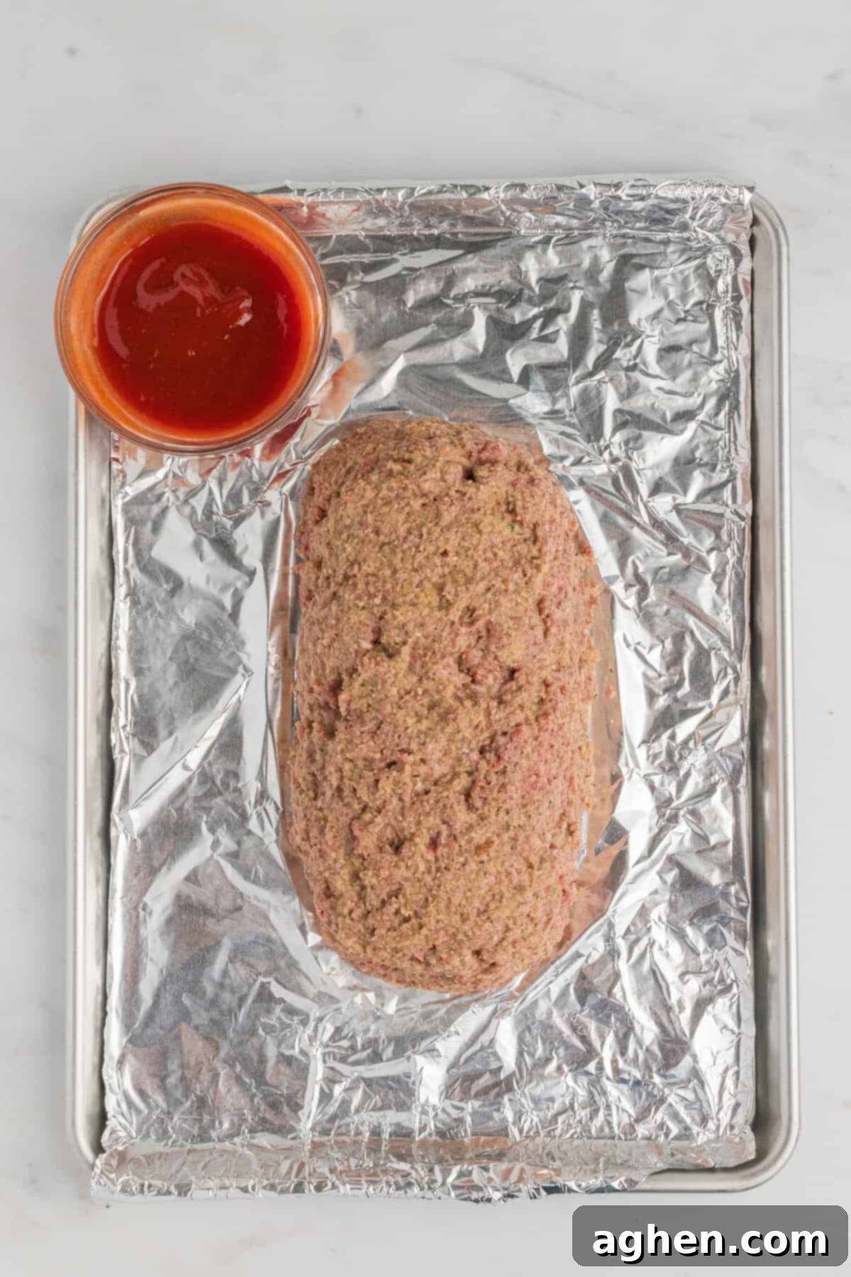 Meatloaf formed into loaf shape on foil lined baking sheet with a bowl of glaze beside it.
