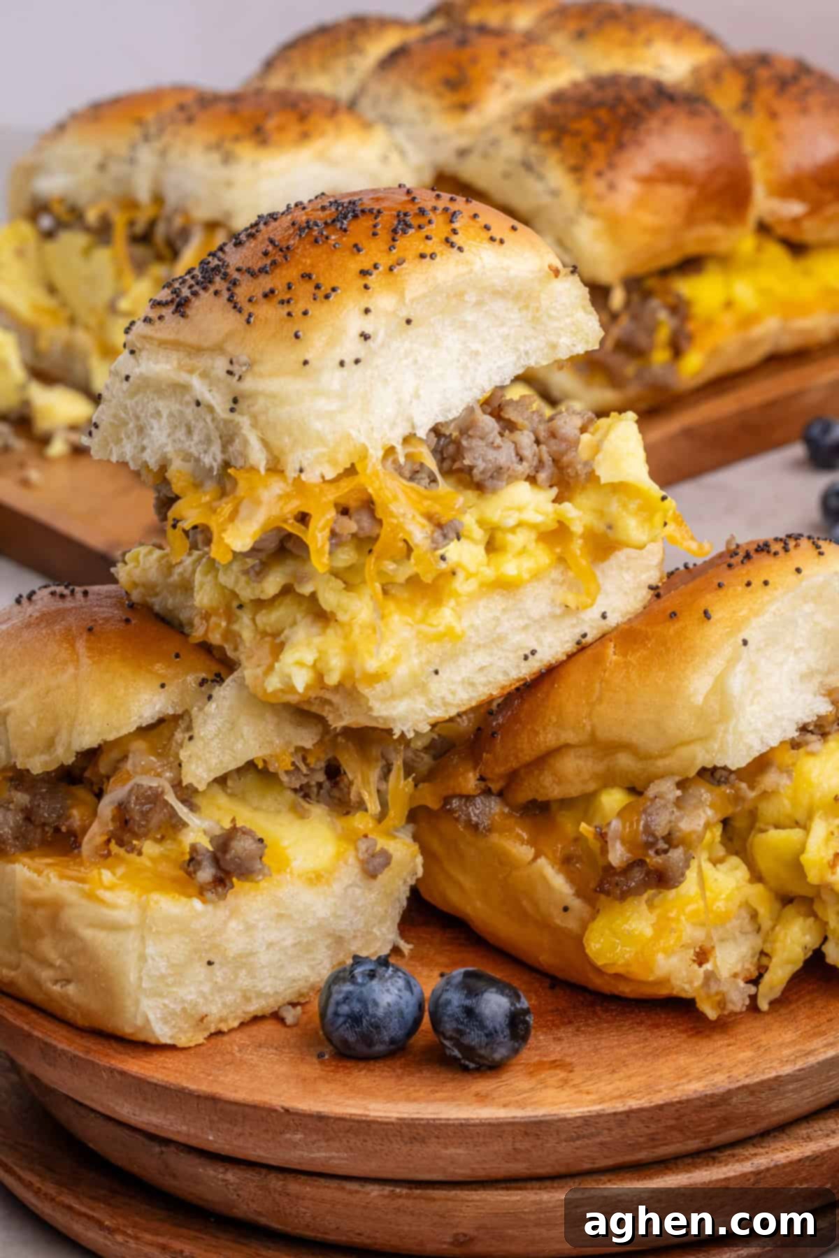 Sweet & Savory Hawaiian Breakfast Bake 2 Delicious sausage, egg, and cheese Hawaiian Roll Breakfast Sliders stacked on a wooden plate with fresh blueberries on the side. More sliders are visible in the background on a serving board, inviting you to grab one.