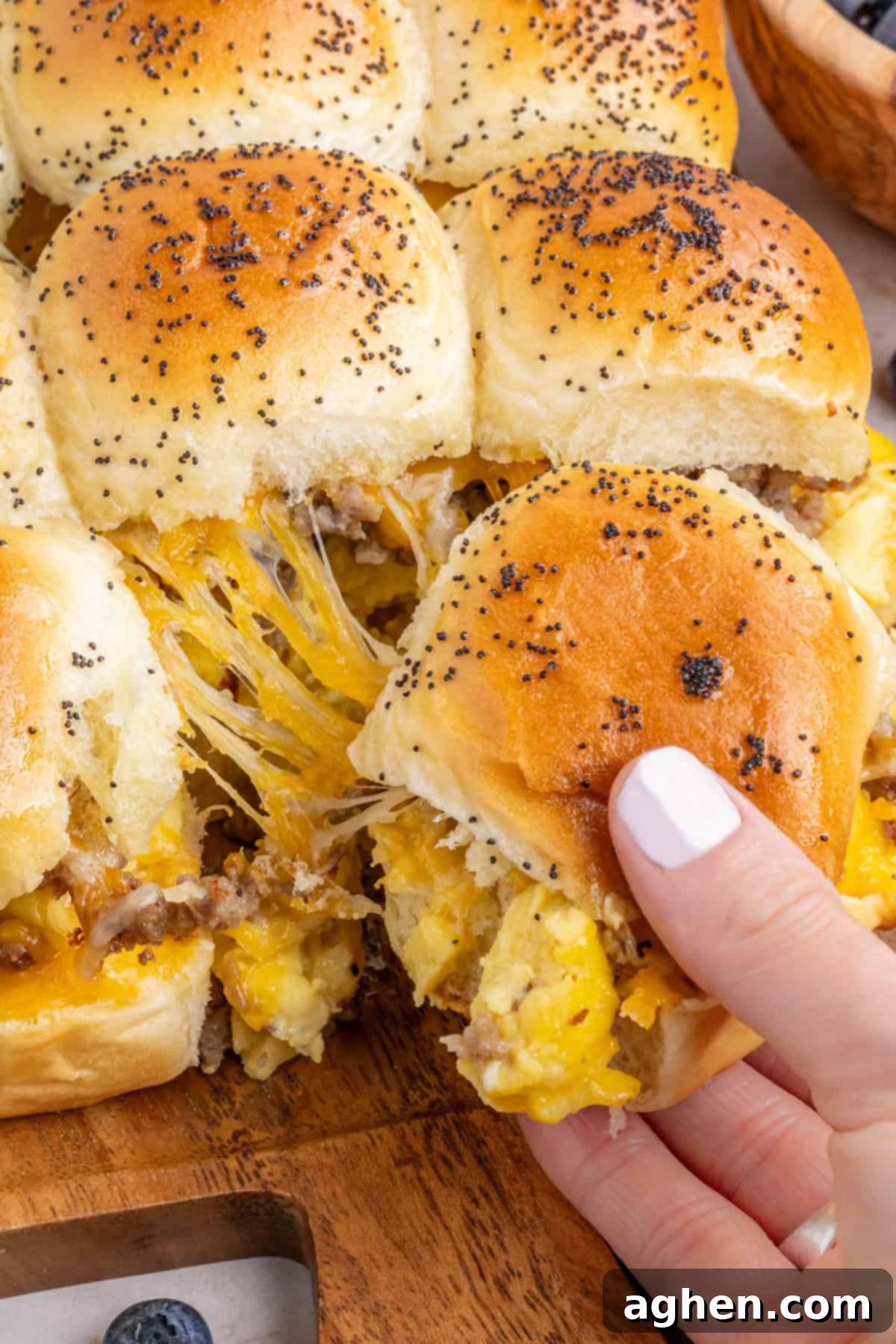 Sweet & Savory Hawaiian Breakfast Bake 3 A hand reaching to pull a single cheesy breakfast slider from a sheet of freshly baked King's Hawaiian breakfast sliders, showcasing the melted cheese and fluffy filling.