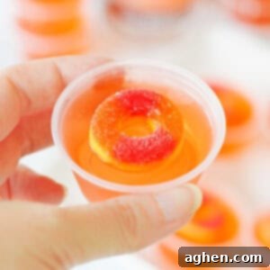 Peach vodka jello shot topped with peach candy ring in a small plastic cup being held by a thumb and forefinger.