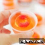 Peach vodka jello shot topped with peach candy ring in a small plastic cup being held by a thumb and forefinger.