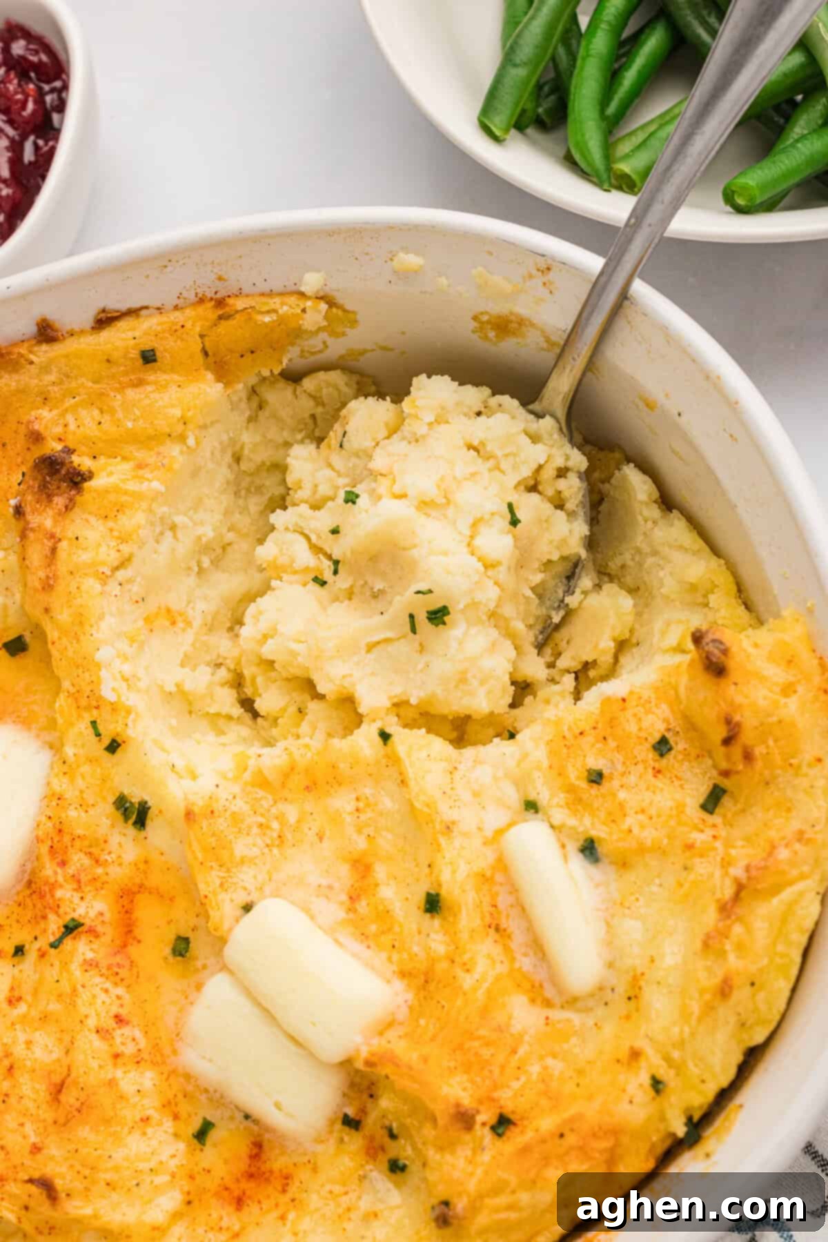 A serving spoon dips into a baking dish filled with golden-topped Party Mashed Potatoes with Cream Cheese and Sour Cream, ready to be dished out.