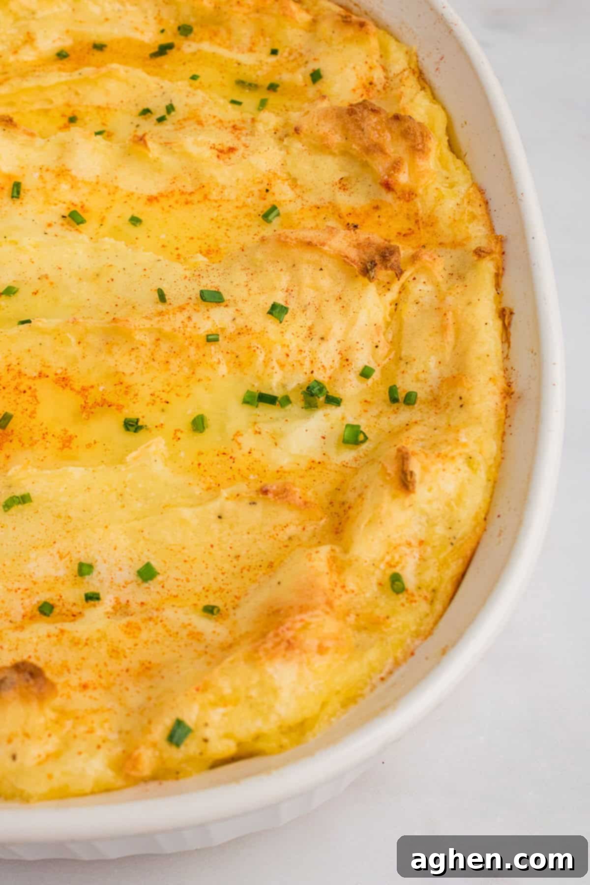 Party mashed potatoes with cream cheese and sour cream baked in a white casserole dish until the top is golden and slightly crispy, garnished with melted butter, paprika, and chives.