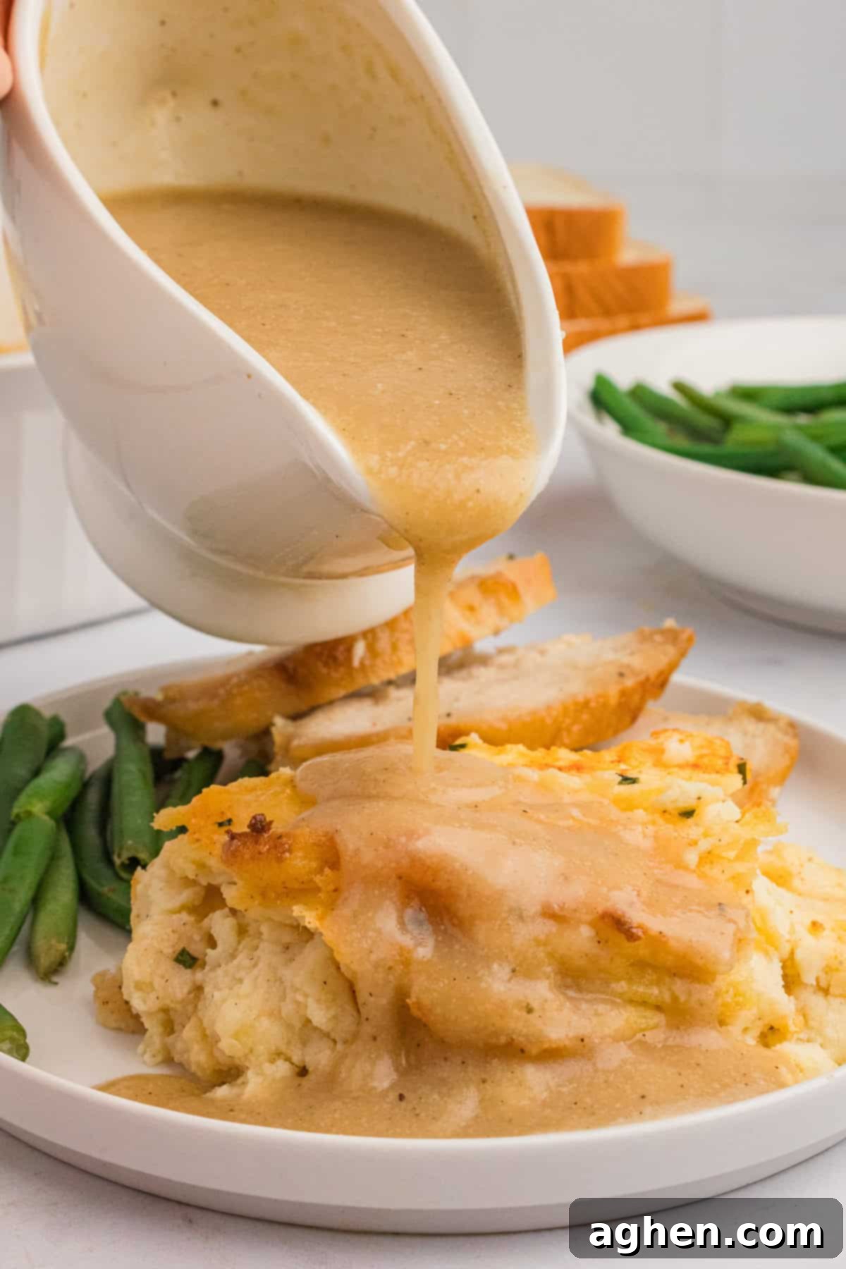 Rich brown gravy is being generously poured over a serving of baked mashed potatoes, nestled on a plate alongside roasted turkey and crisp green beans, creating a mouth-watering holiday meal.