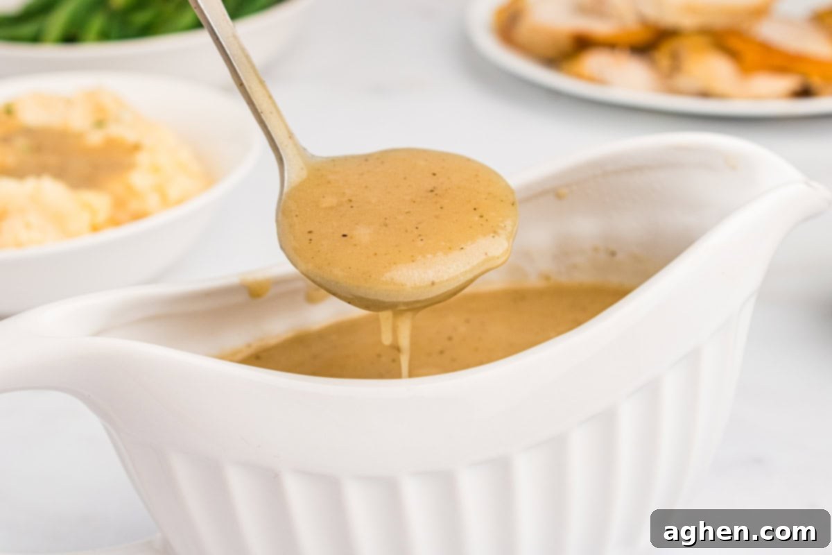 Easy homemade turkey gravy, presented beautifully in a white gravy boat, with some spooned into a ladle. In the background, a spread of roast turkey, creamy mashed potatoes, and vibrant green beans creates a mouth-watering scene.