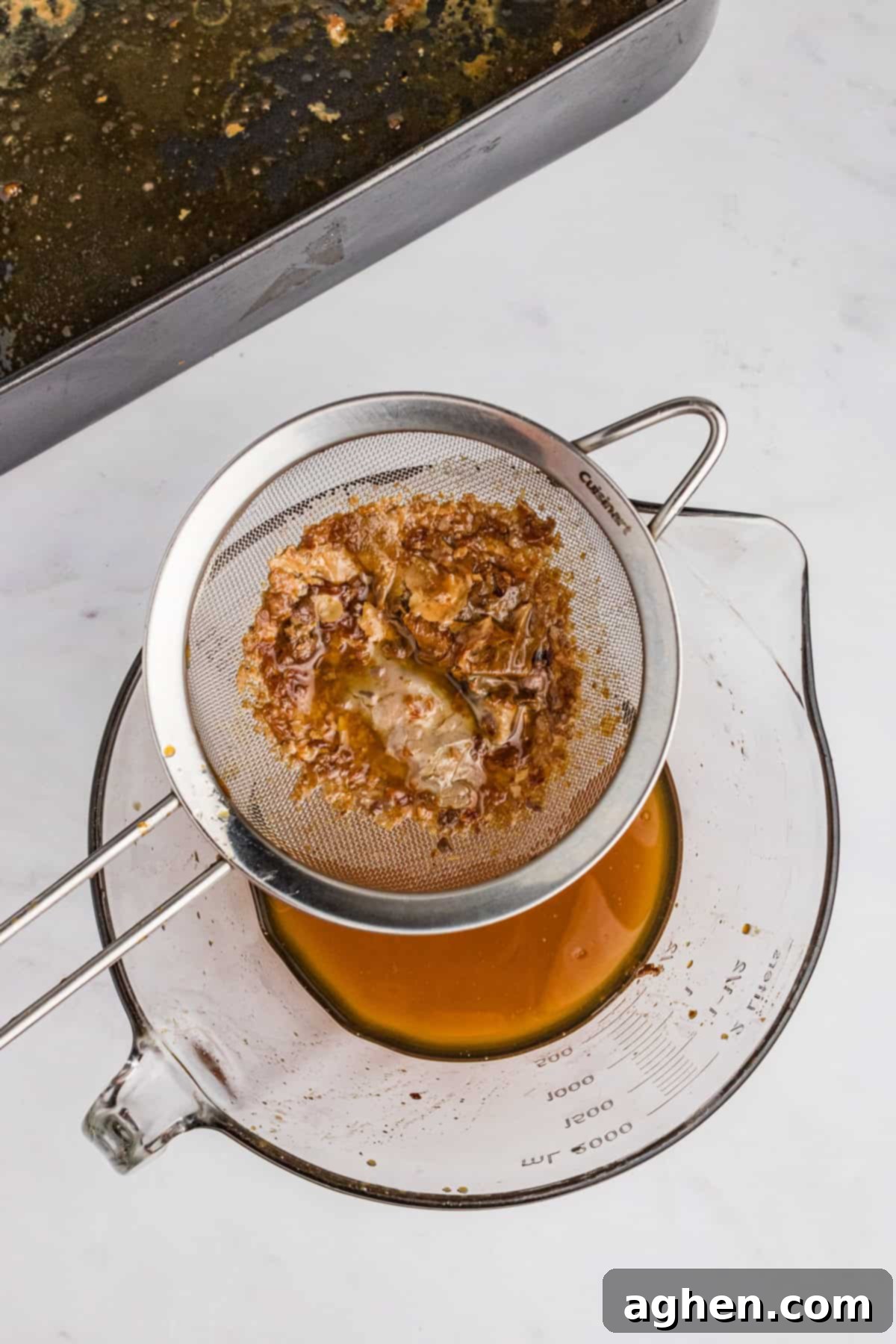 Pan drippings being poured through a strainer into a large glass bowl with a spout.