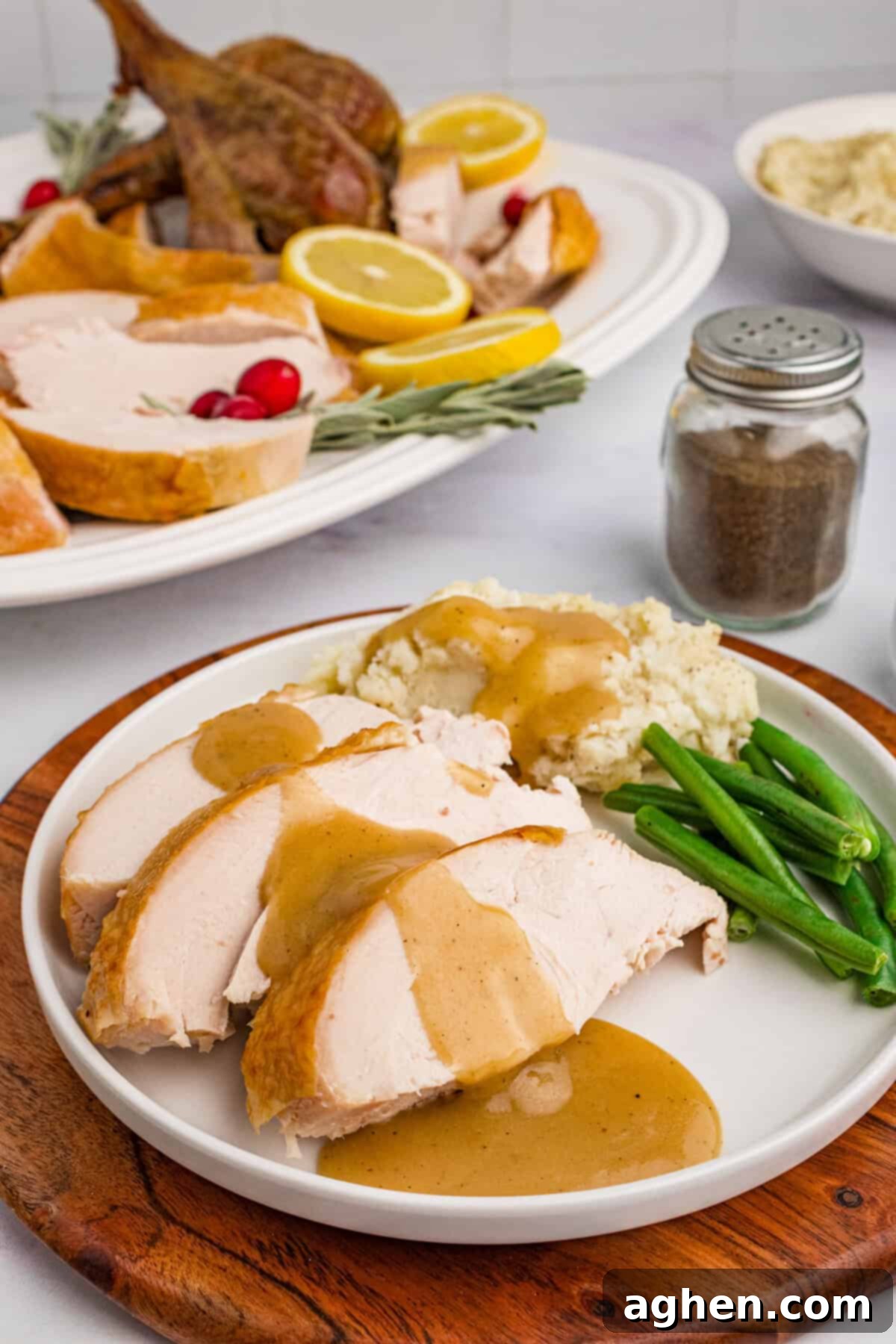 Sliced turkey with gravy on a plate alongside green beans and mashed potatoes, with roasted turkey and mashed potatoes in the background.