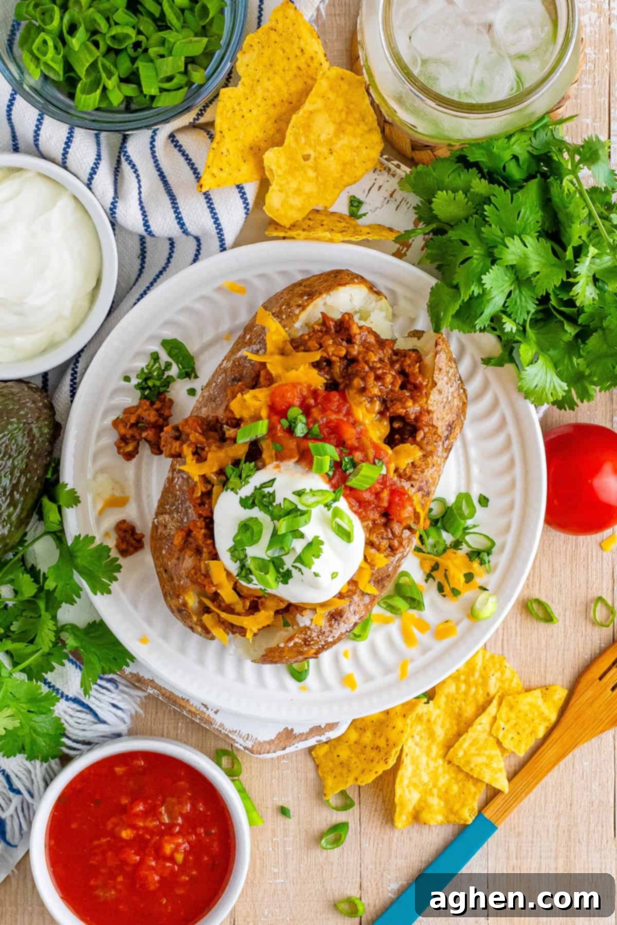A top-down view of a fully loaded taco baked potato, generously topped with seasoned ground beef, melted cheese, fresh salsa, sour cream, and vibrant green onions. It is surrounded by additional taco toppings and tortilla chips on a rustic wooden table.