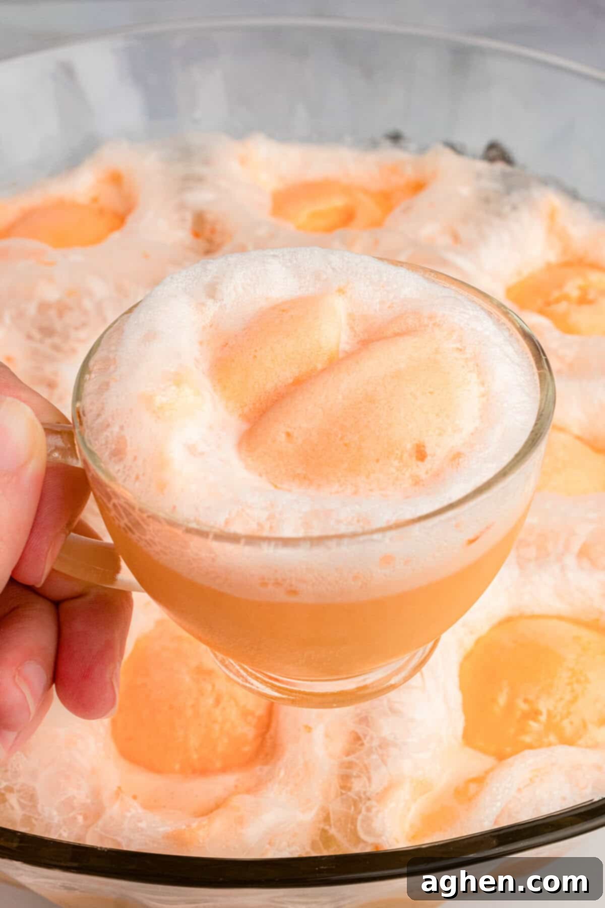 Hand holding glass of champagne punch topped with scoop of sherbet with the rest of the sherbet punch in a large bowl behind it.