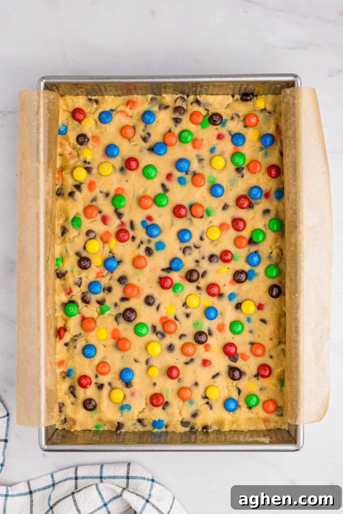 Chocolate chip M&M cookie dough topped with extra M&M's, pressed into a parchment-lined 9 x 13 inch baking pan, ready for the oven.