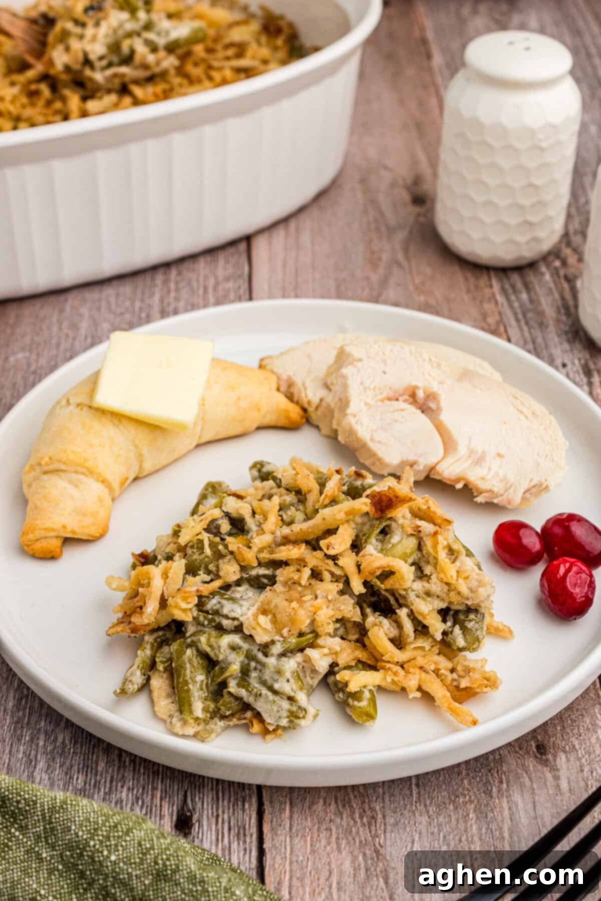 A serving of green bean casserole with crispy fried onions plated alongside sliced turkey, vibrant cranberry sauce, and a golden, buttered crescent roll, ready for a festive meal.