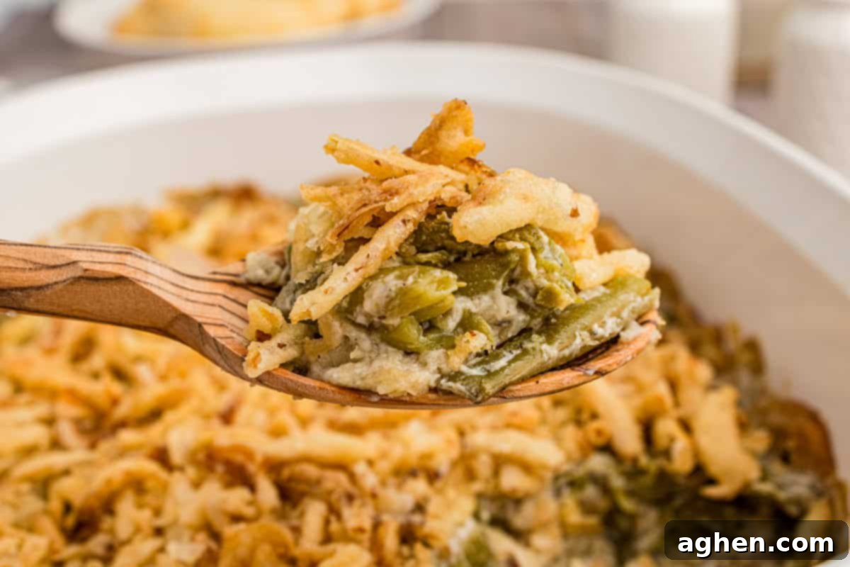 Classic green bean casserole generously topped with crispy fried onions, with a wooden spoon poised to remove a warm, creamy portion from the baking dish.