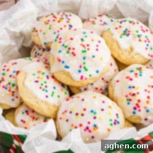 A close-up of Italian ricotta cookies topped with smooth white icing and colorful round sprinkles, arranged in a festive green tin lined with parchment paper. The cookies are soft, fluffy, and stacked to showcase their homemade charm.