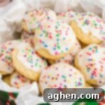 A close-up of Italian ricotta cookies topped with smooth white icing and colorful round sprinkles, arranged in a festive green tin lined with parchment paper. The cookies are soft, fluffy, and stacked to showcase their homemade charm.