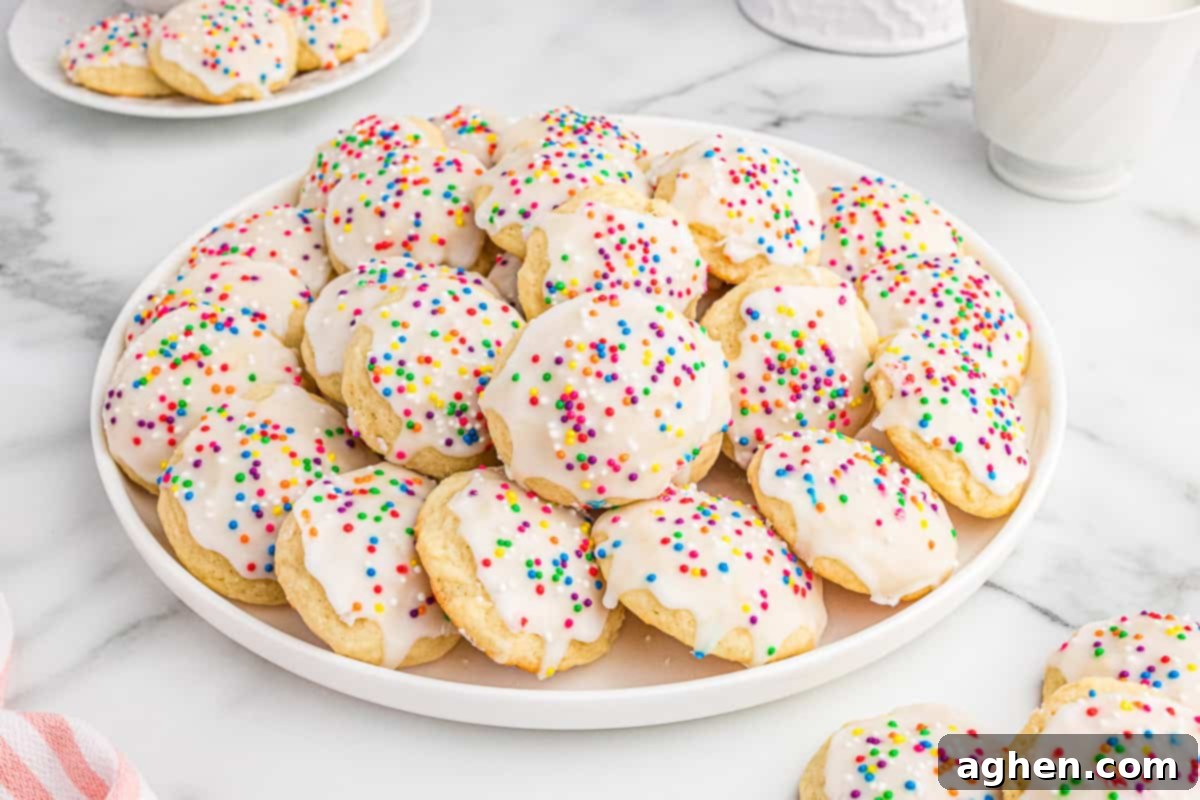 A white plate piled high with Italian ricotta cookies topped with smooth white icing and colorful sprinkles. Additional cookies are visible on a smaller plate in the background, set on a marble surface, creating a bright and festive display.